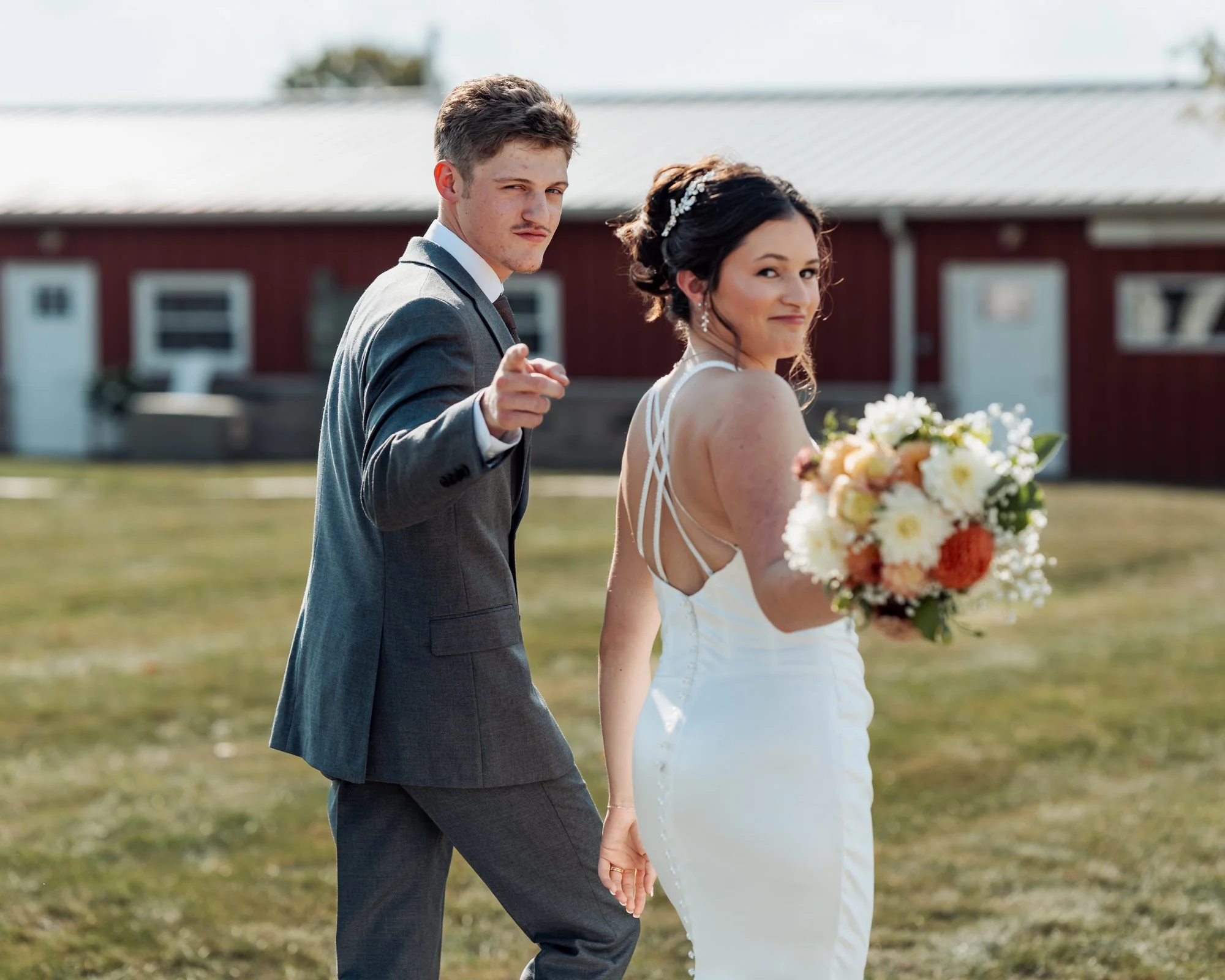 A newlywed couple walking outdoors, the groom points at the camera, and the bride holds a bouquet, with a barn in the background.