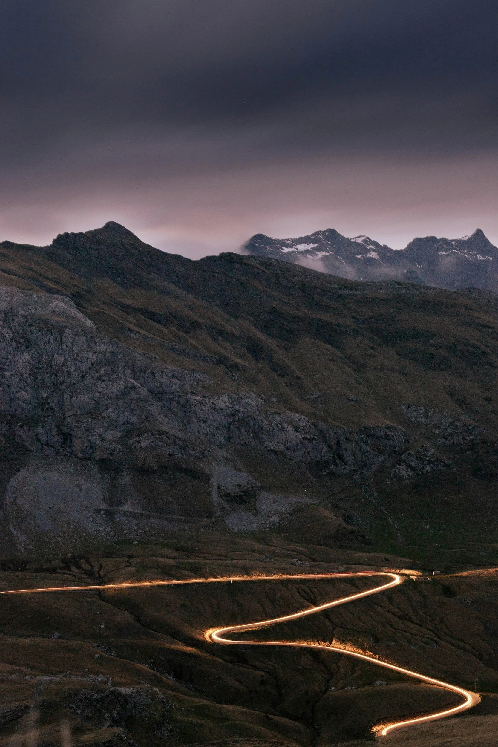 A mountain landscape at dusk with a winding dirt road illuminated by car lights leading through the valley to the mountains in the background.
