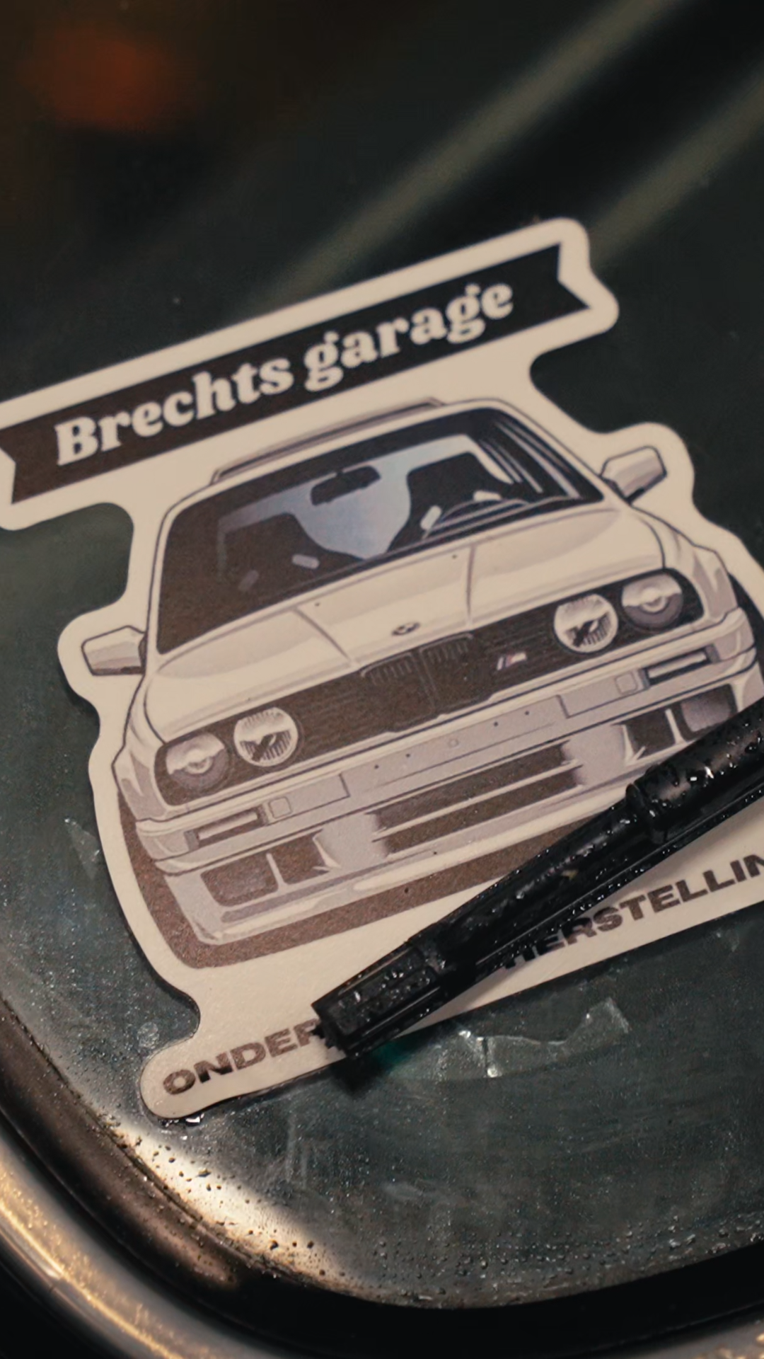 A sticker of a classic car with the text 'Brecht's garage' above it, placed on a black surface with a small part of a black object nearby.