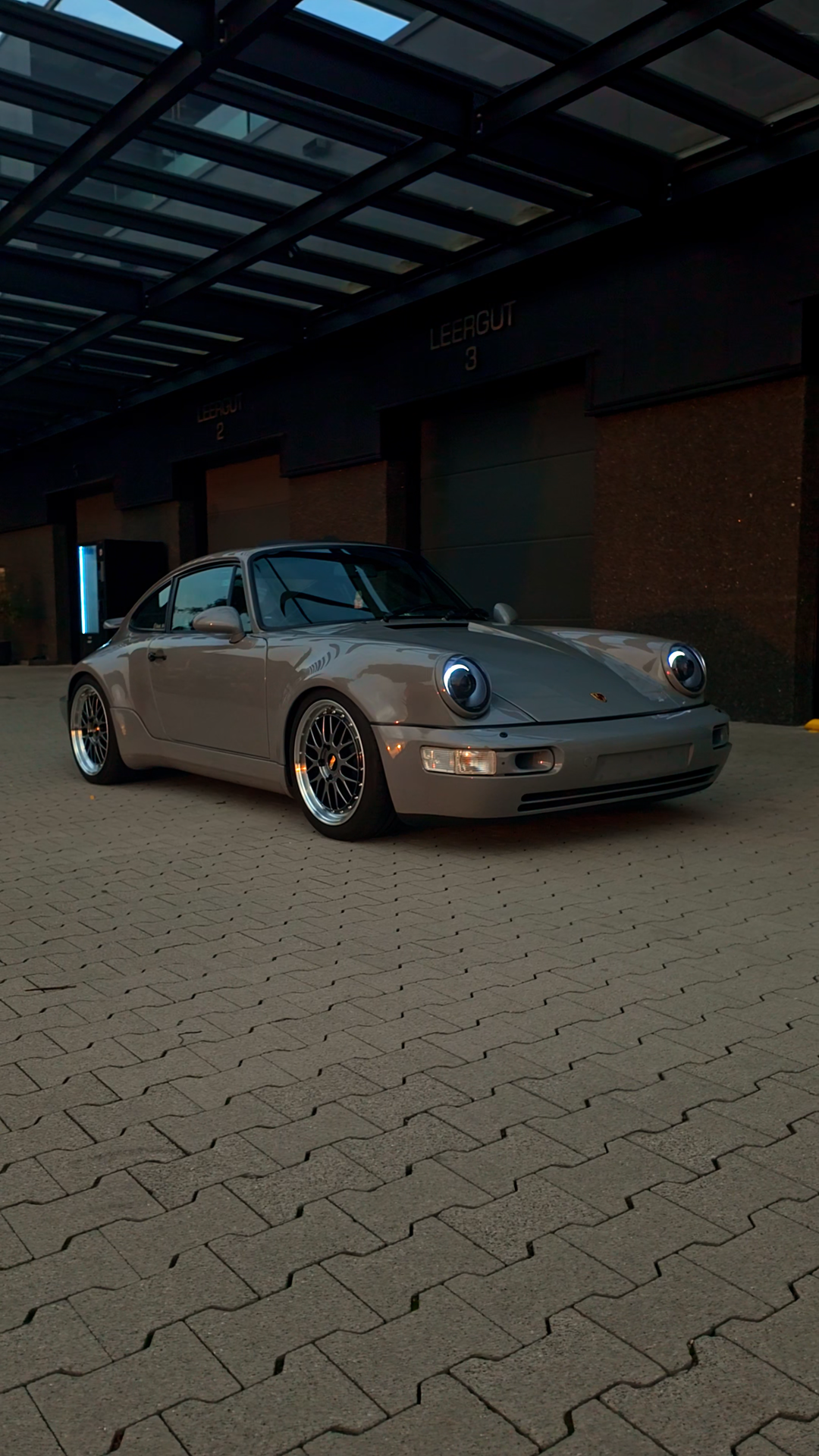 A silver Porsche 911 parked outside a building with numbered garage doors numbered 2 and 3 under a glass canopy.