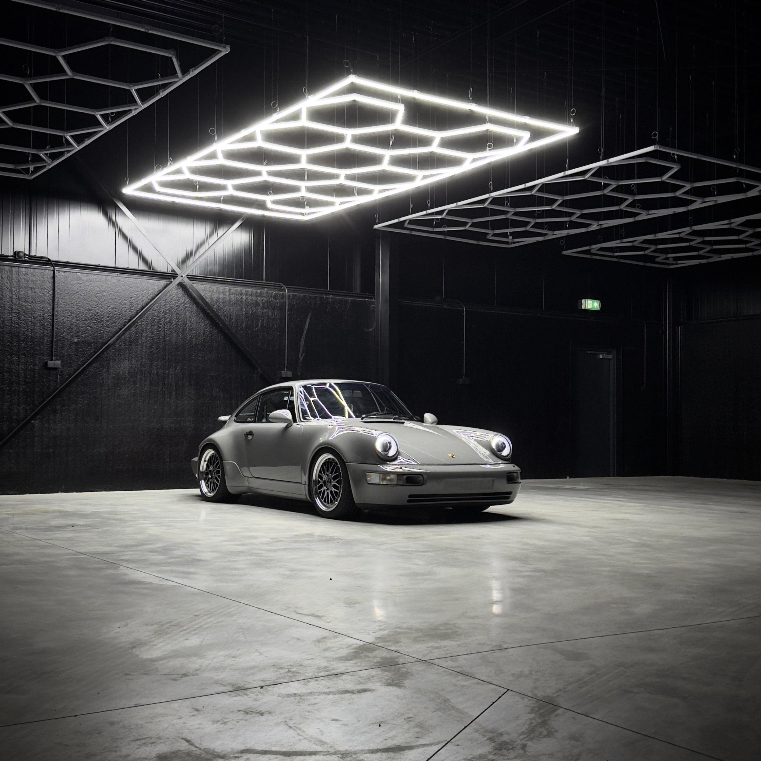 A vintage silver Porsche 911 sports car parked inside a dark, modern showroom with geometric LED light fixtures on the ceiling.