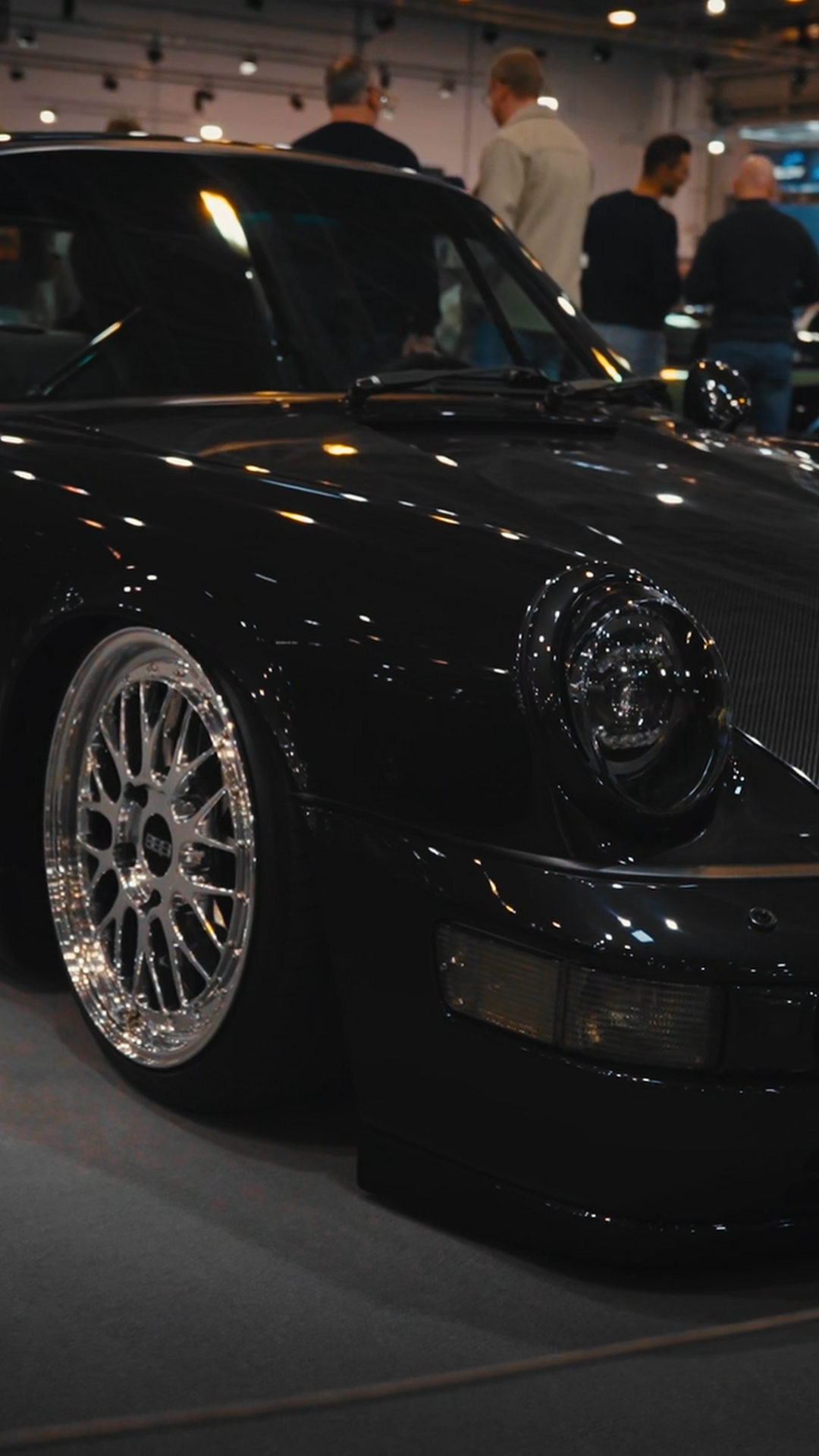 Close-up of a black sports car with shiny wheels on display at an indoor car show, with people visible in the background.