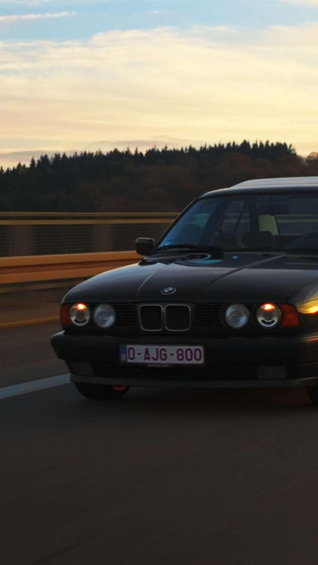 A black vintage BMW car driving on a highway during sunset with a forested landscape in the background.