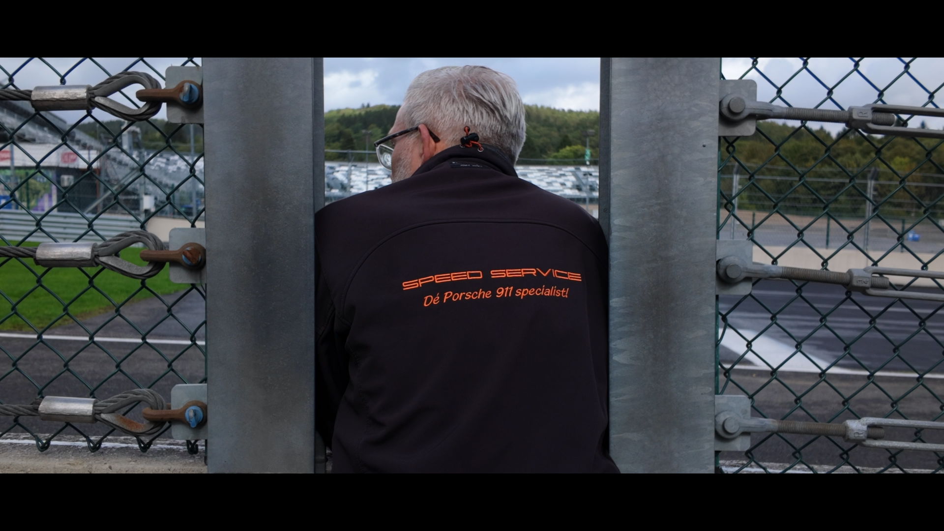 A man with gray hair wearing a black jacket with orange text that reads 'Speed Service Porsche 911 specialist' leans against a chain-link fence at a race track, looking away from the camera.