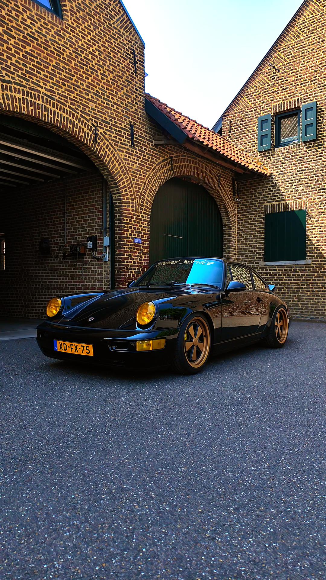 A black vintage Porsche sports car with gold rims parked in front of a brick building with arched doorways and windows with blue shutters.