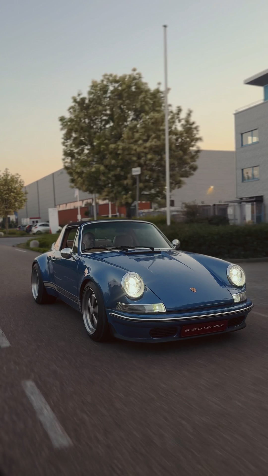 A vintage blue Porsche 911 driving on a city street at sunset, with a driver and passenger visible inside.