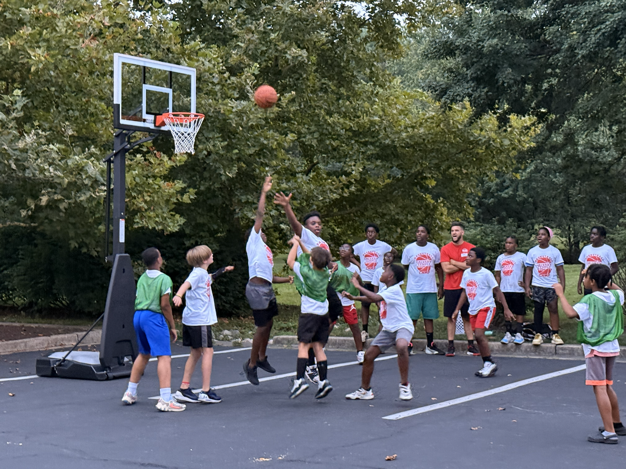 Children and teenagers playing basketball on an outdoor court surrounded by trees during daytime.