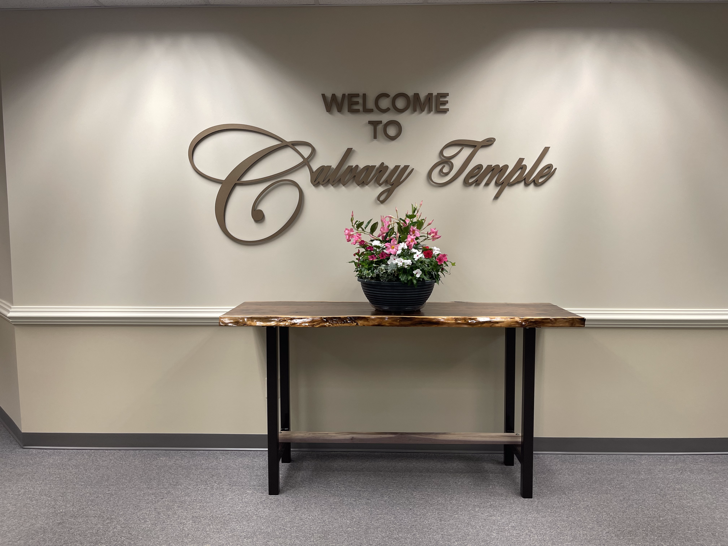 Indoor scene showing a wooden table with a flower arrangement in a black pot, against a wall with the words 'Welcome to Calvary Temple' in large decorative lettering.