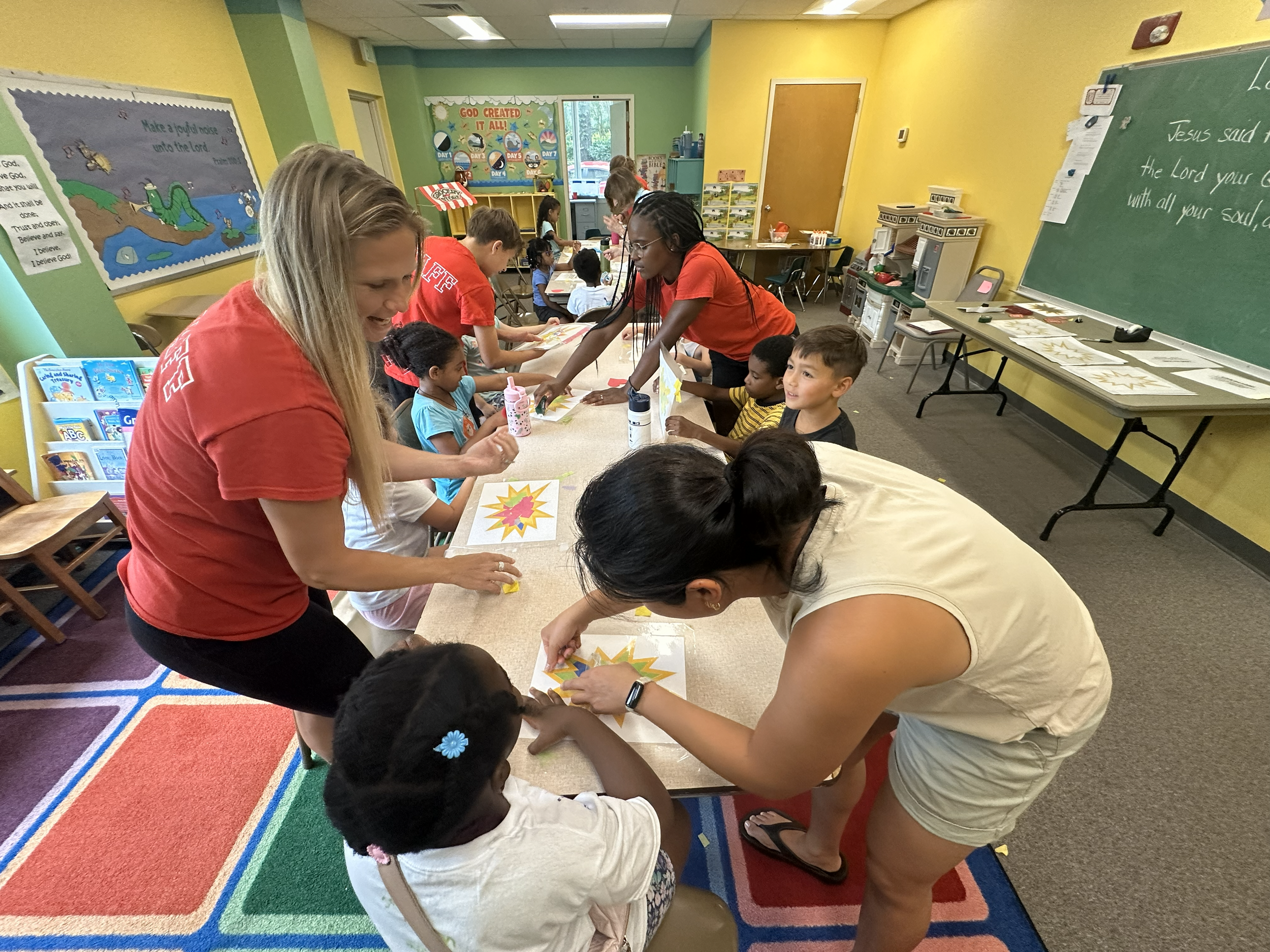 Children and adults gathered around a table in a colorful classroom, working on craft projects with bright paper and supplies, with educational posters and a chalkboard in the background.