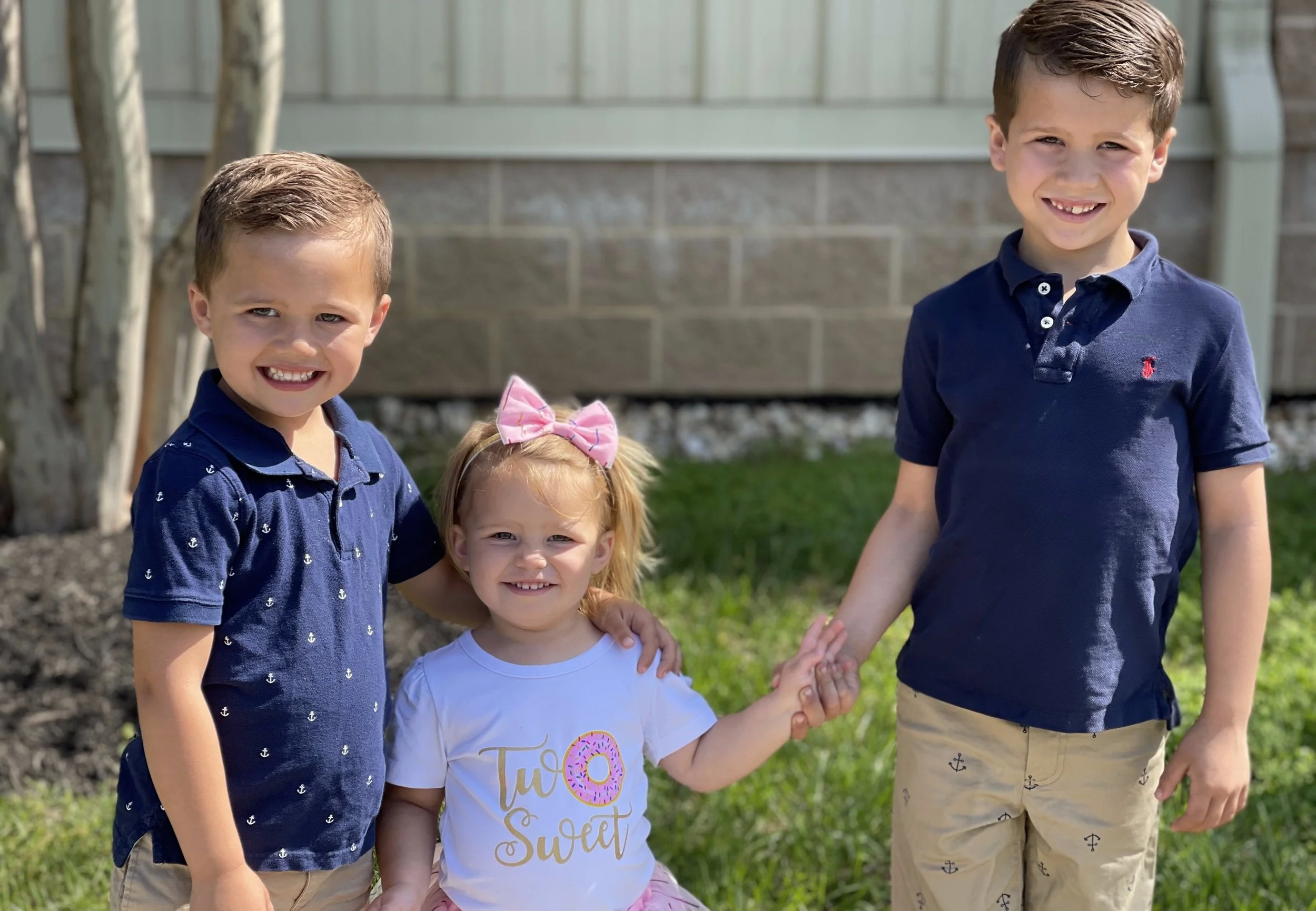 Three children, two boys and a girl, standing outdoors on grass in front of a , smiling at the camera. The girl has a pink bow in her hair and wears a white shirt with a pink donut graphic that says 'Two Sweet'. The boy on the left has short brown hair, wears a navy blue polo shirt with small white anchor patterns, and beige shorts. The boy on the right has short brown hair, wears a plain navy blue polo shirt with a red logo, and beige shorts with small anchor patterns.