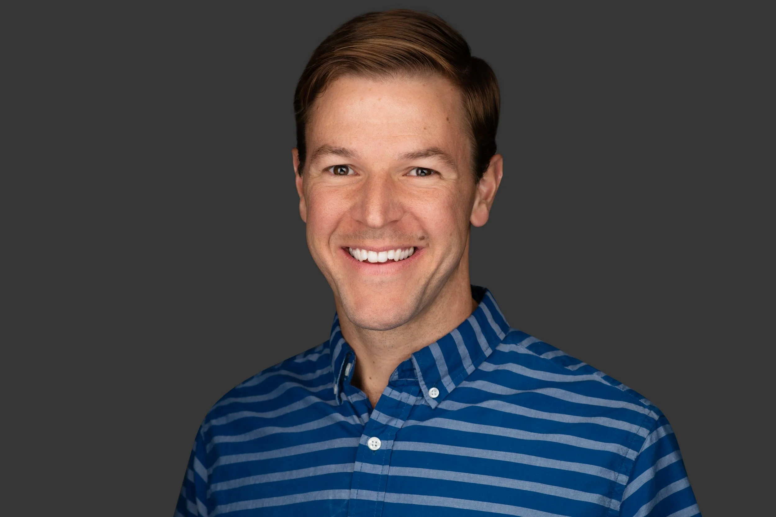 Headshot of a smiling Pastor with short brown hair, wearing a blue and gray striped button-up shirt, against a dark gray background.