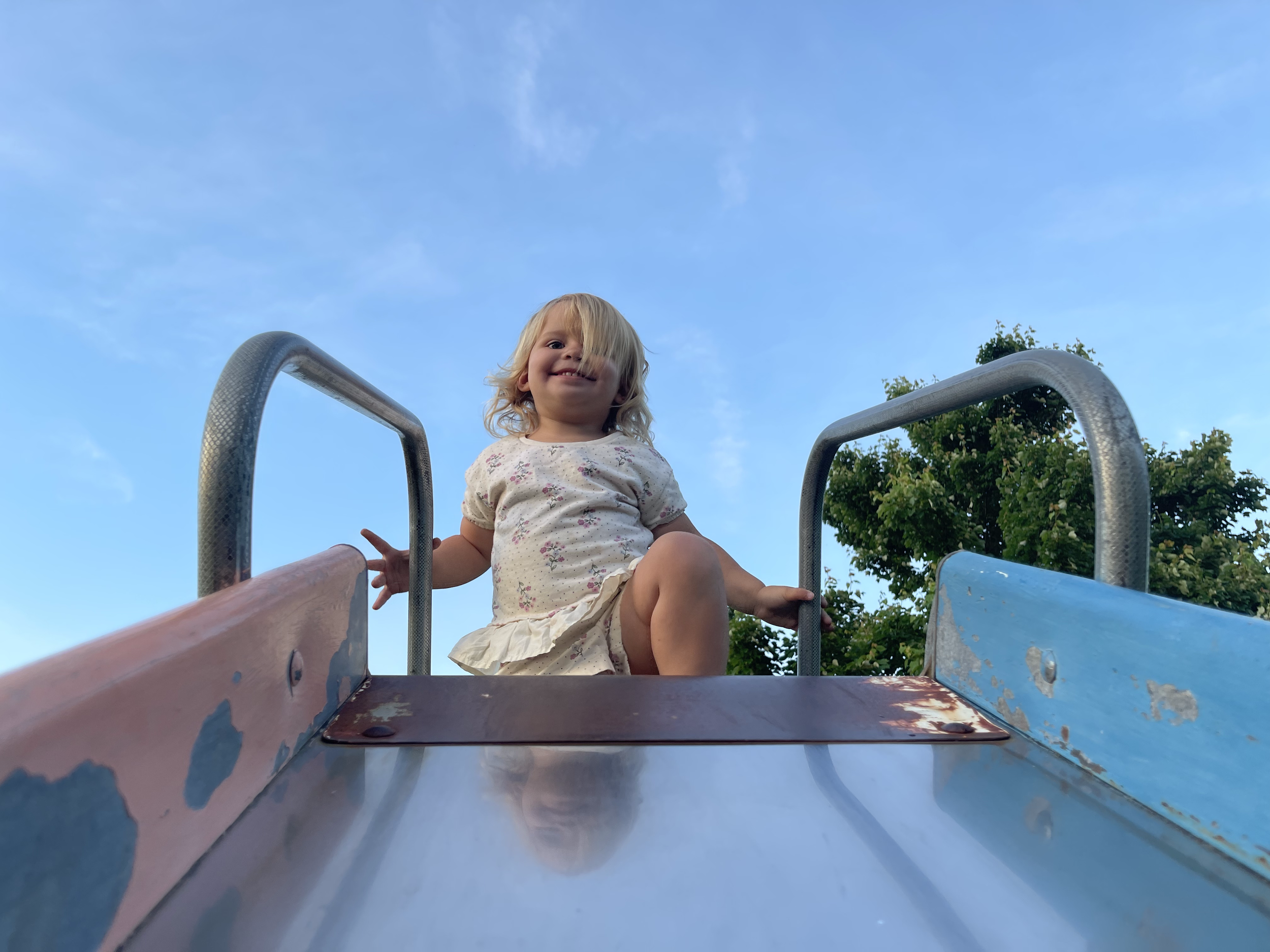 A young girl with blonde hair smiling while sitting at the top of a playground slide, with a clear blue sky and green trees in the background.