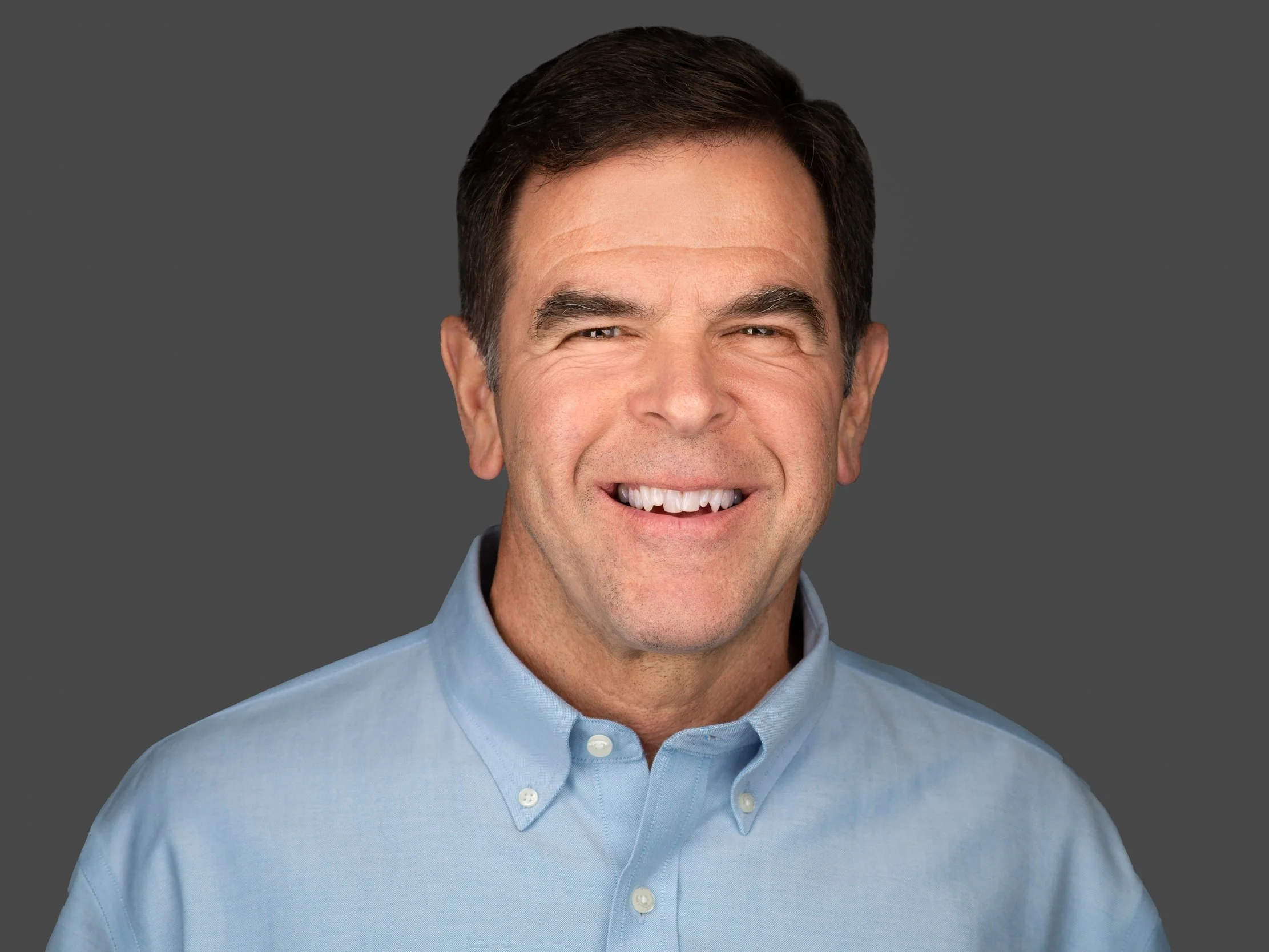 Close-up photo of a smiling Pastor with dark hair, wearing a light blue button-up shirt, against a solid gray background.