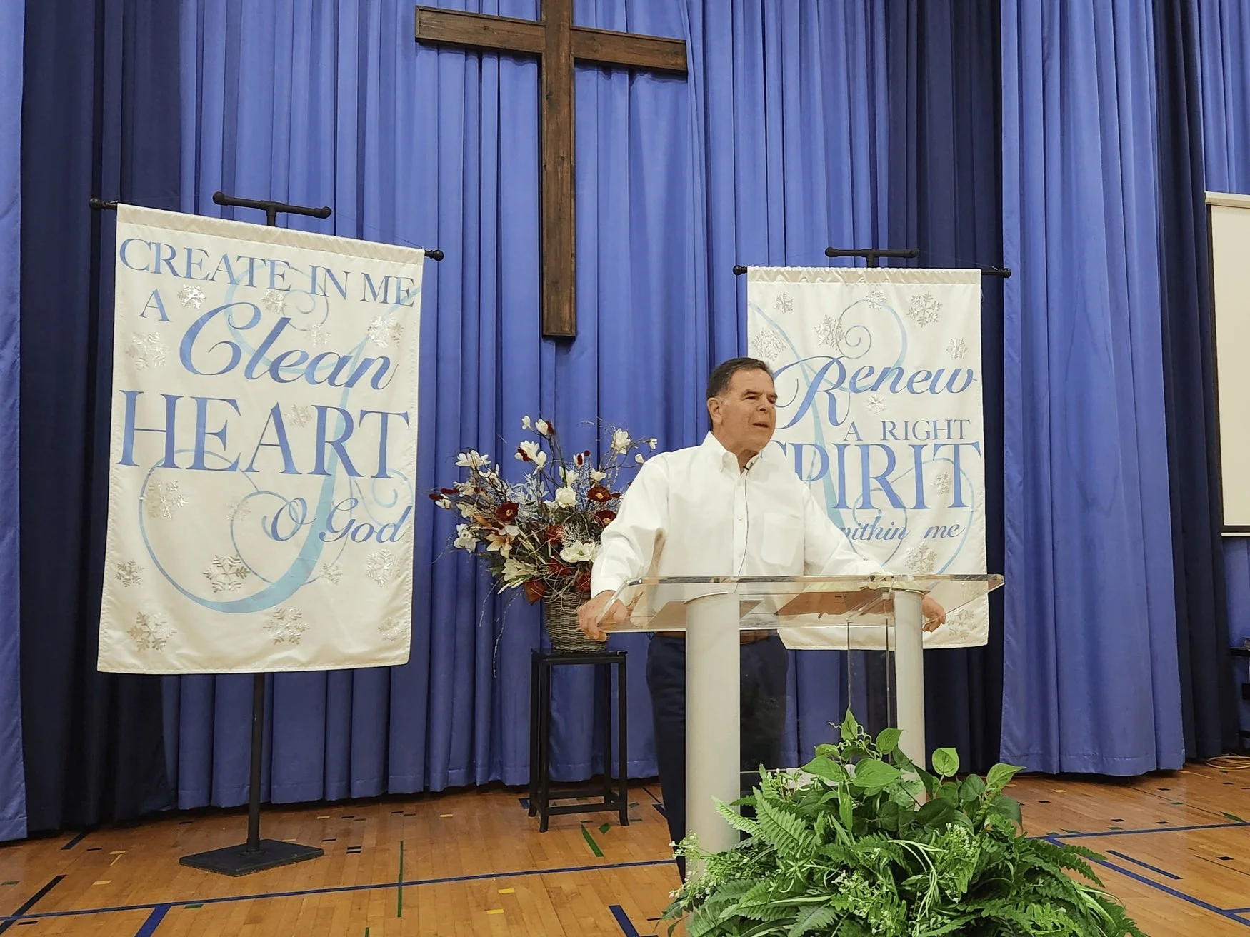 A Pastor standing behind a clear podium, speaking in front of blue curtains with two banners, surrounded by plants and flowers, with a cross mounted on the wall behind him.