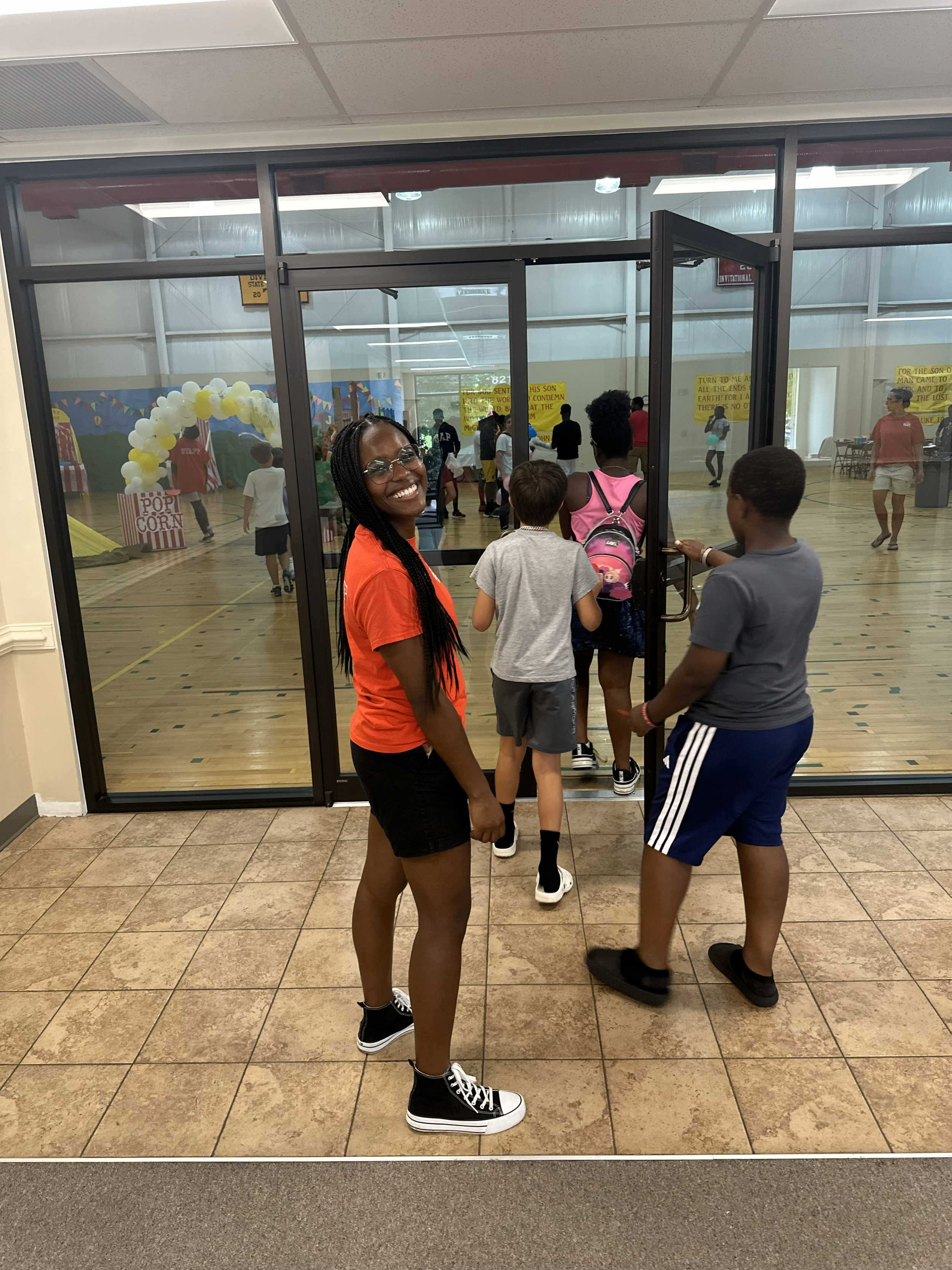 A young woman with long braided hair and glasses, smiling, stands outside a gymnasium entrance as children walk in for an event. The gymnasium has a wooden floor, colorful decorations, and themed signs for a carnival or festival.