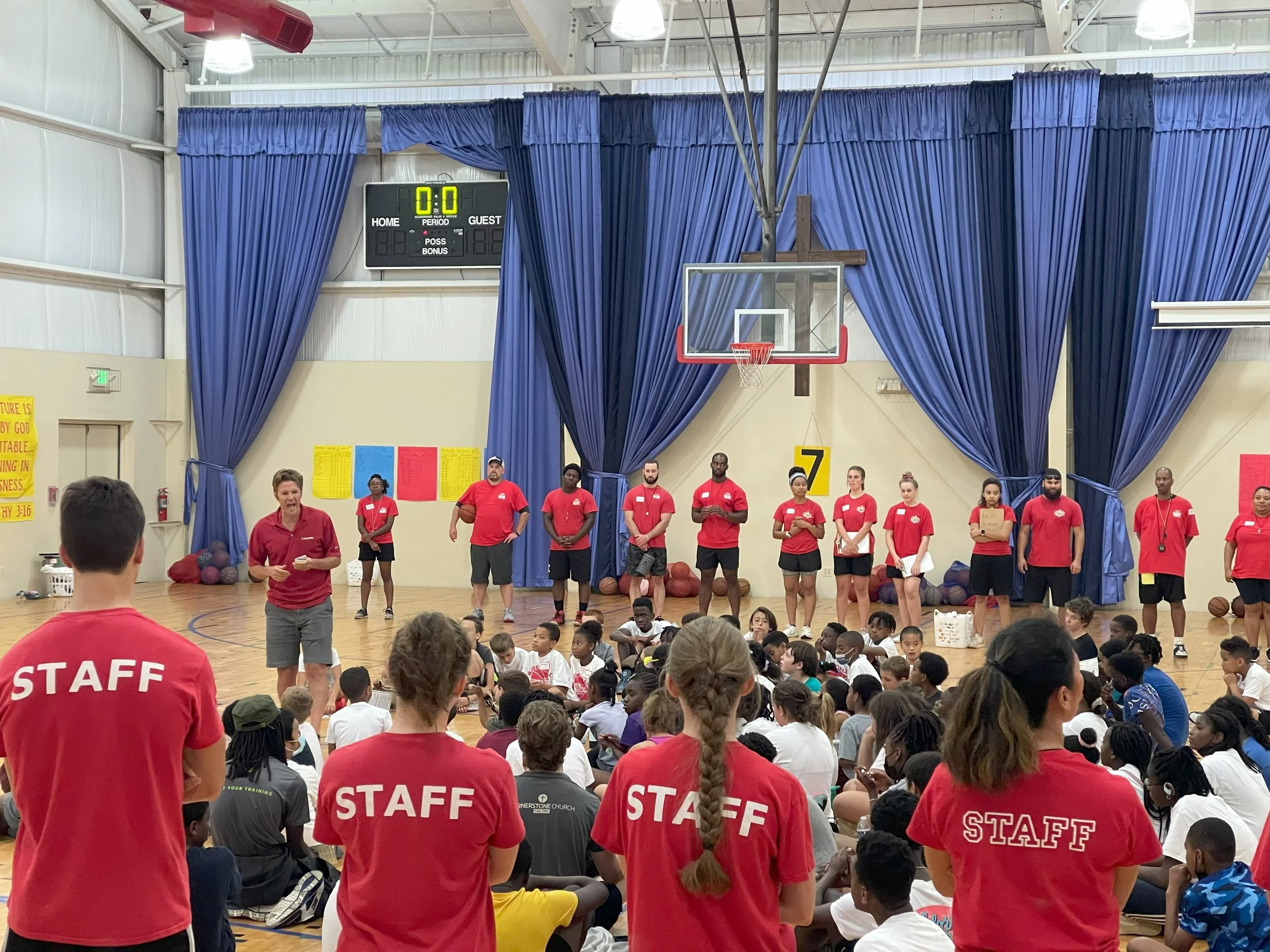 A large gymnasium with a basketball court filled with children seated on the floor, facing a group of staff members and coaches standing at the front near a basketball hoop. The staff members wear red shirts with