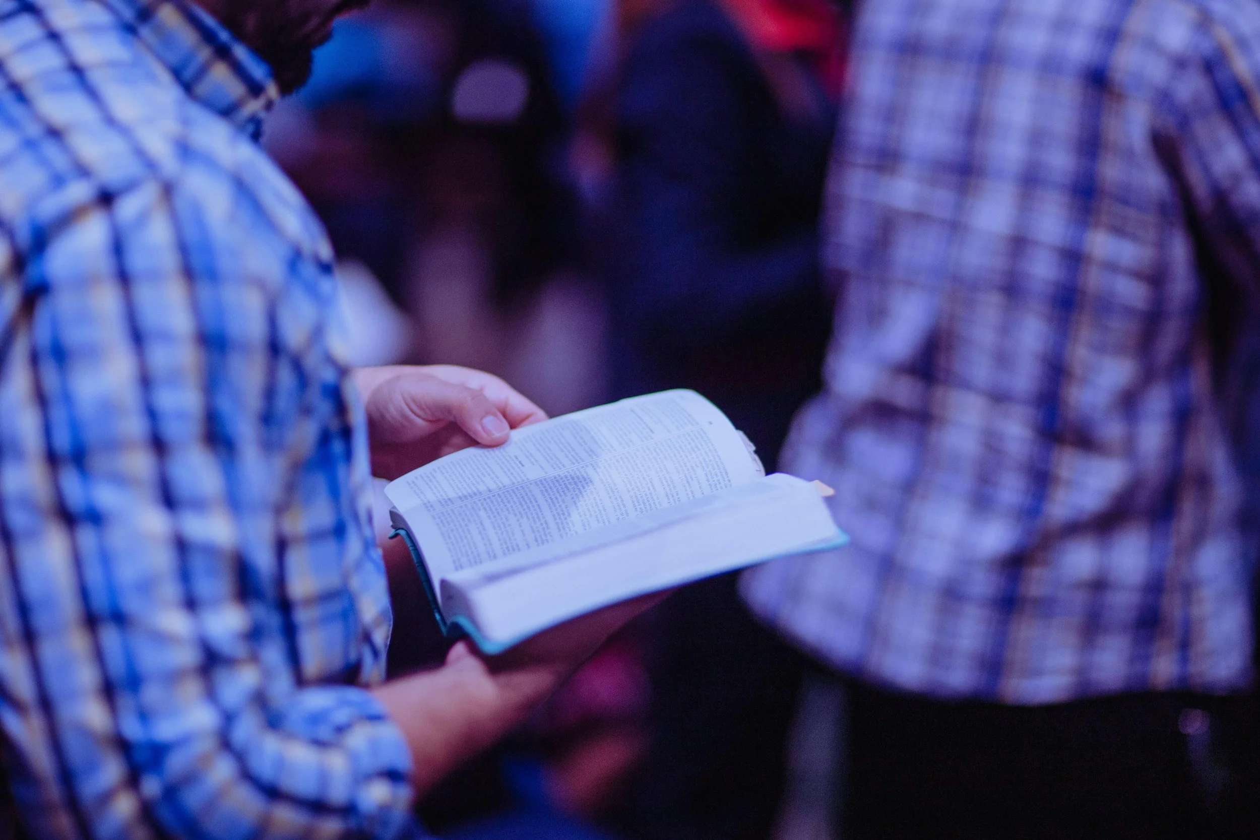 Person holding an open Bible or book in a dimly lit environment, with others around.
