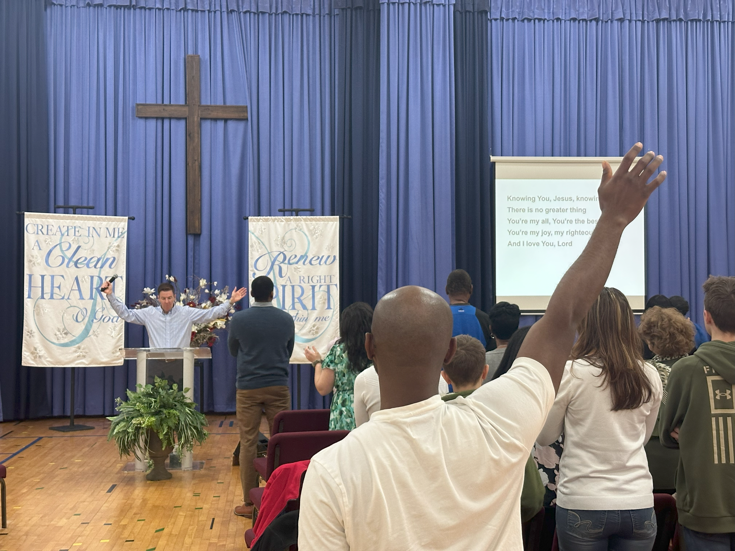 A man leads a prayer at the front of a church with a cross on a blue curtain backdrop. Several people stand and raise their hands. A large screen displays lyrics relating to Jesus and worship. Decorative banners read encouraging messages about the Holy Spirit and the heart.