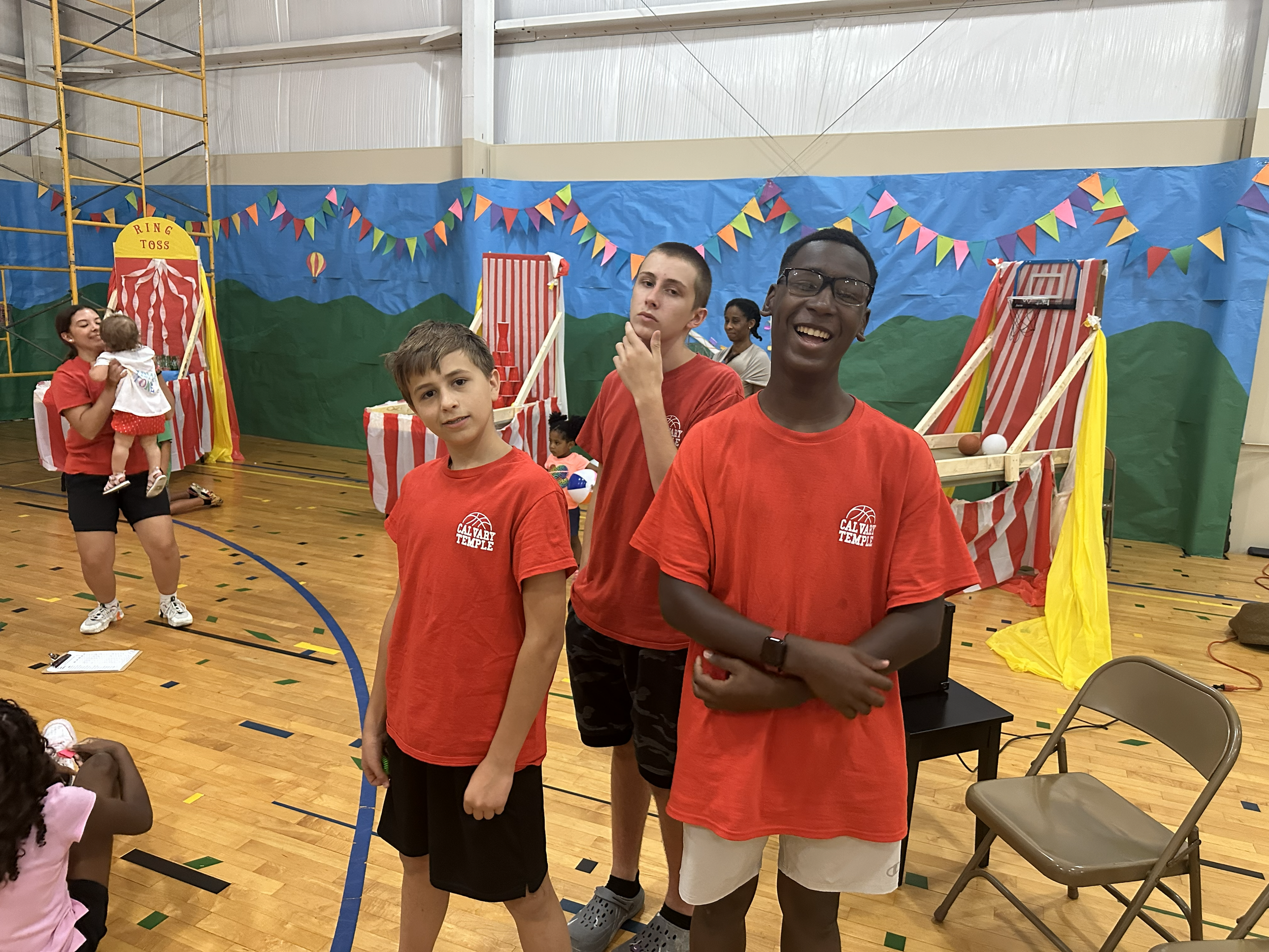 Group of children at a carnival-style event inside a gymnasium with colorful decorations, including a ring toss game, and children playing in the background.