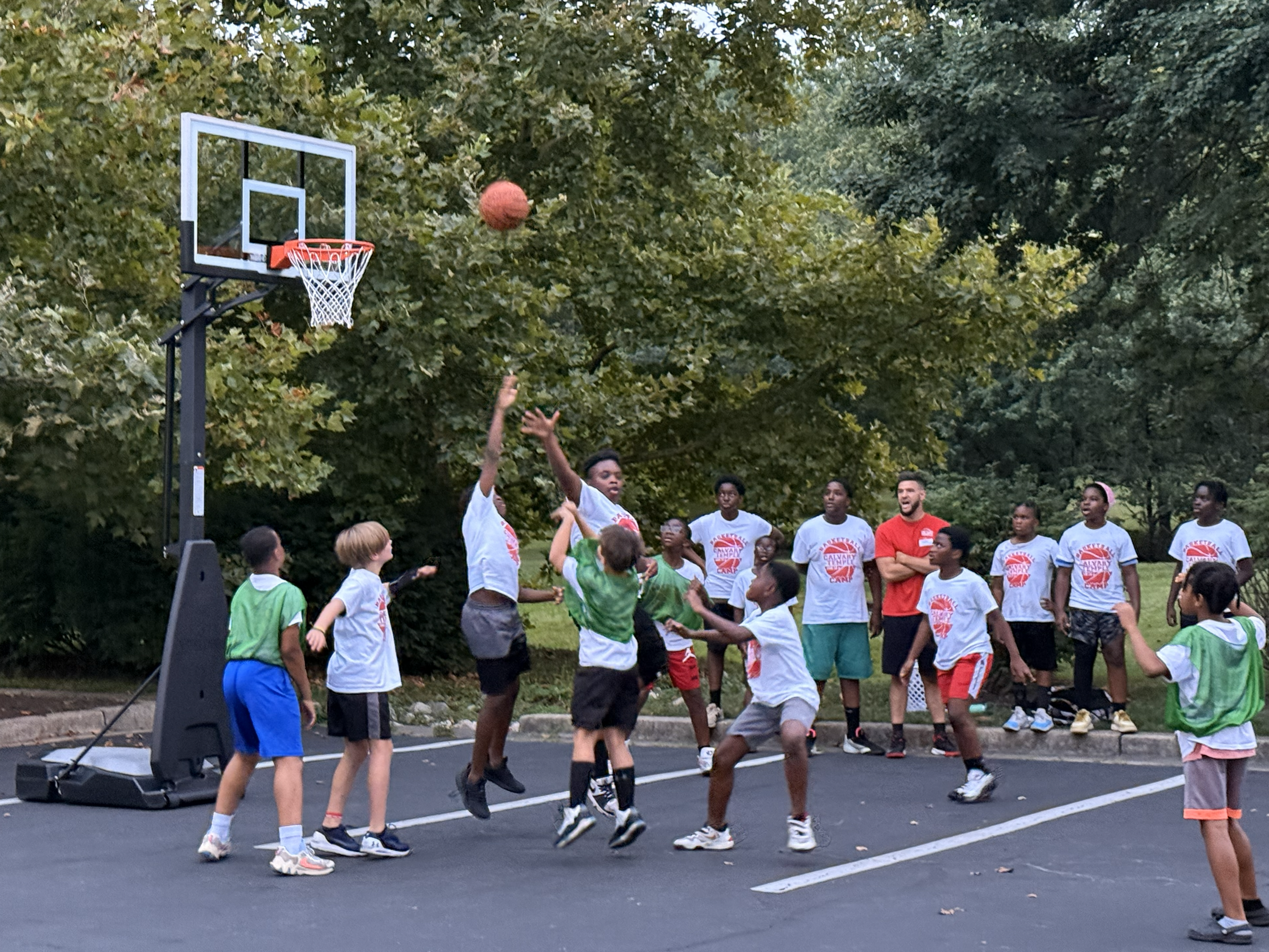 Children playing basketball outdoors with onlookers in loin, surrounded by trees.