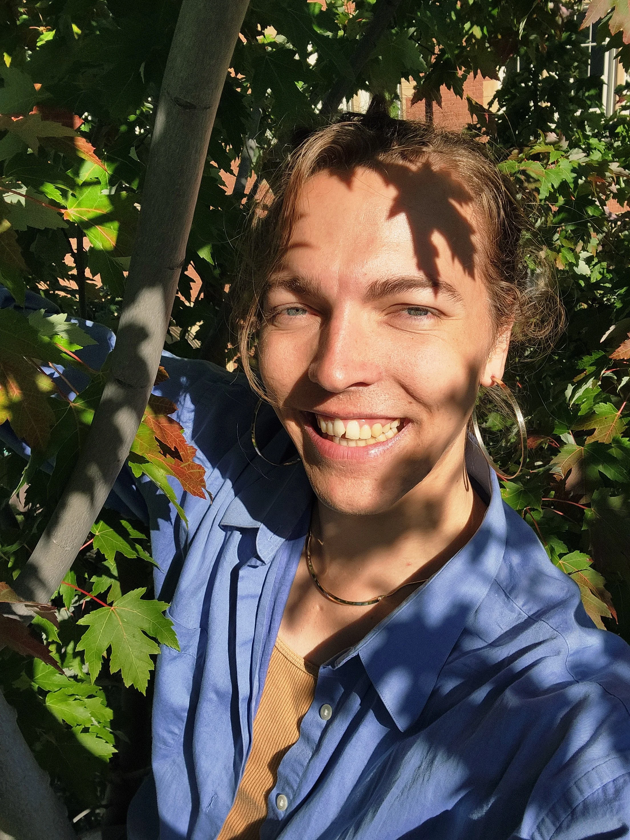 Person smiling outdoors wearing a blue bike helmet and large hexagonal sunglasses, standing in front of a wooden railing and green trees with a body of water in the background.