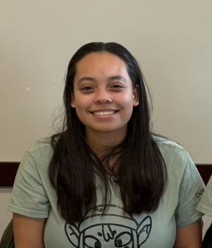 A woman with long brown hair and a cheerful smile is taking a selfie in a car with her golden retriever dog sitting behind her. The dog is looking at the camera with its tongue slightly out.