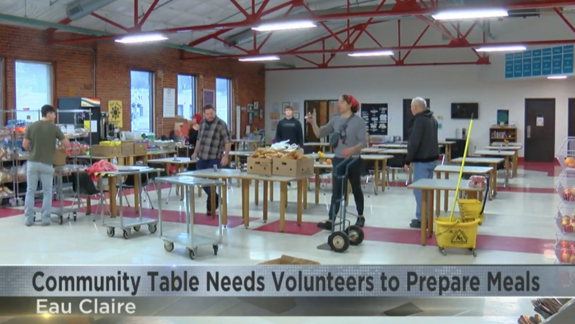 Inside a community center, volunteers are preparing meals on tables, with some people organizing food items and others cleaning. There are cleaning supplies and food supplies around, and the caption reads 'Community Table Needs Volunteers to Prepare Meals' with the location 'Eau Claire'.
