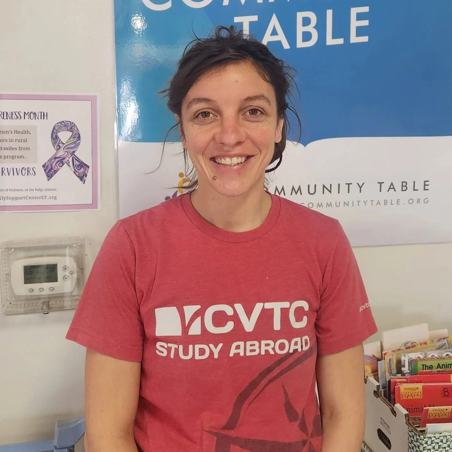 A woman smiling, wearing a red Study Abroad T-shirt, standing in front of a blue and white sign for Community Table.