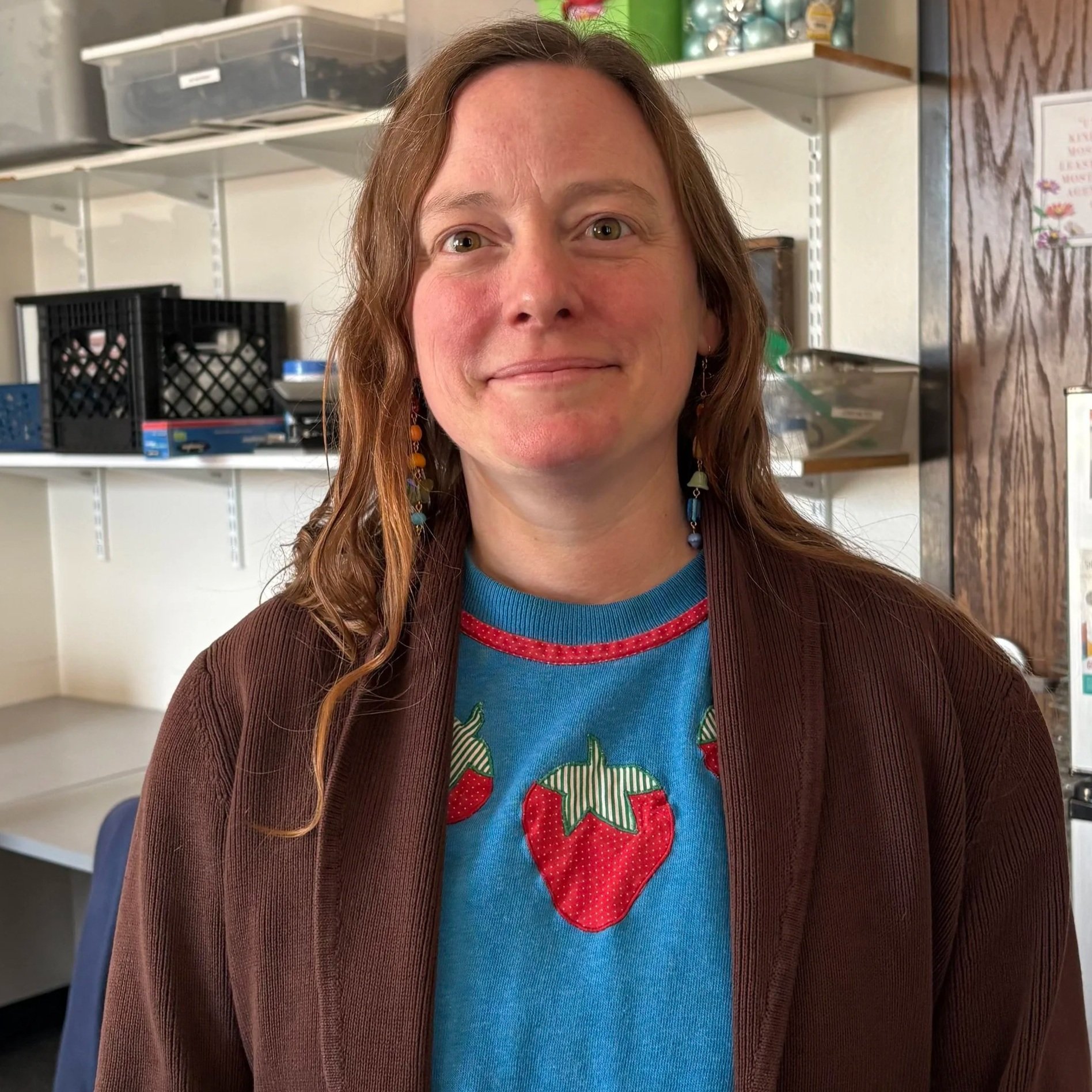 A woman with long wavy hair wearing a blue shirt with strawberry print and a brown jacket, standing in a room with shelves filled with storage boxes and containers.