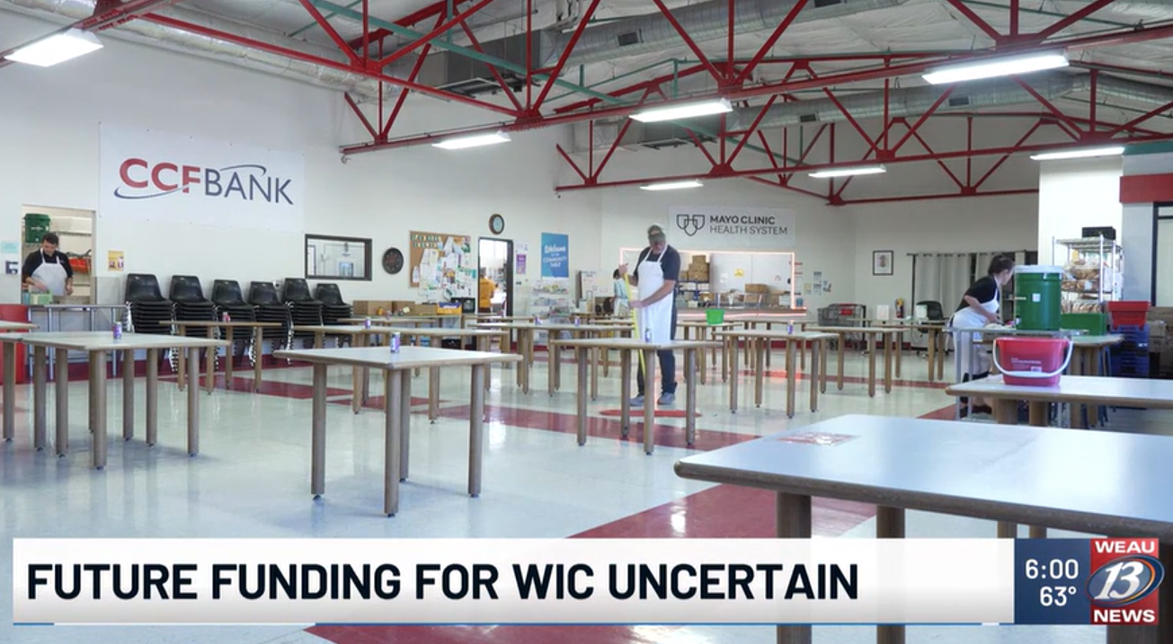 Empty dining area with several tables and chairs, some stacks of chairs, a bulletin board, and staff preparing food or packages at the Mayo Clinic food system. The room features high ceilings with red steel beams, and a news banner at the bottom reads, 'Future funding for WIC uncertain' with a time, temperature, and news station logo.