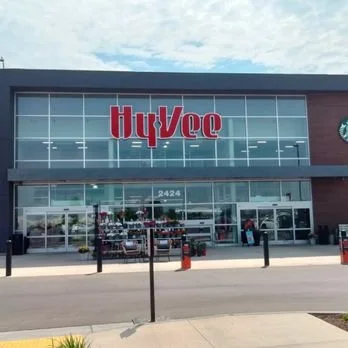 Exterior of a Hy-Vee grocery store with large glass windows and entrance doors, storefront signage, and shopping carts outside.