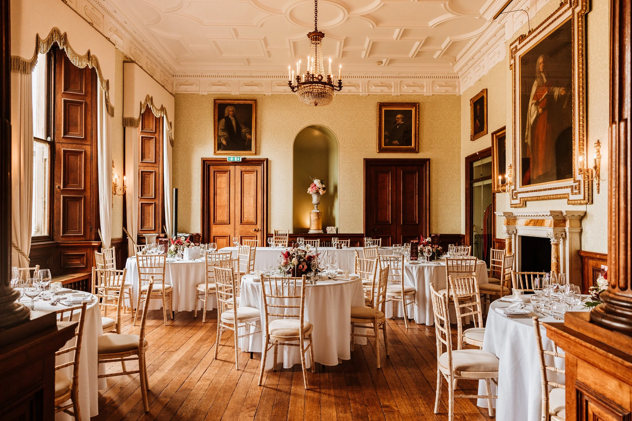 Elegant dining room with round tables covered in white tablecloths, wooden chairs, floral centerpieces, and large windows with curtains. The room has a chandelier, portrait paintings, a fireplace, and wooden paneling at Weston Hall, Staffordshire. 