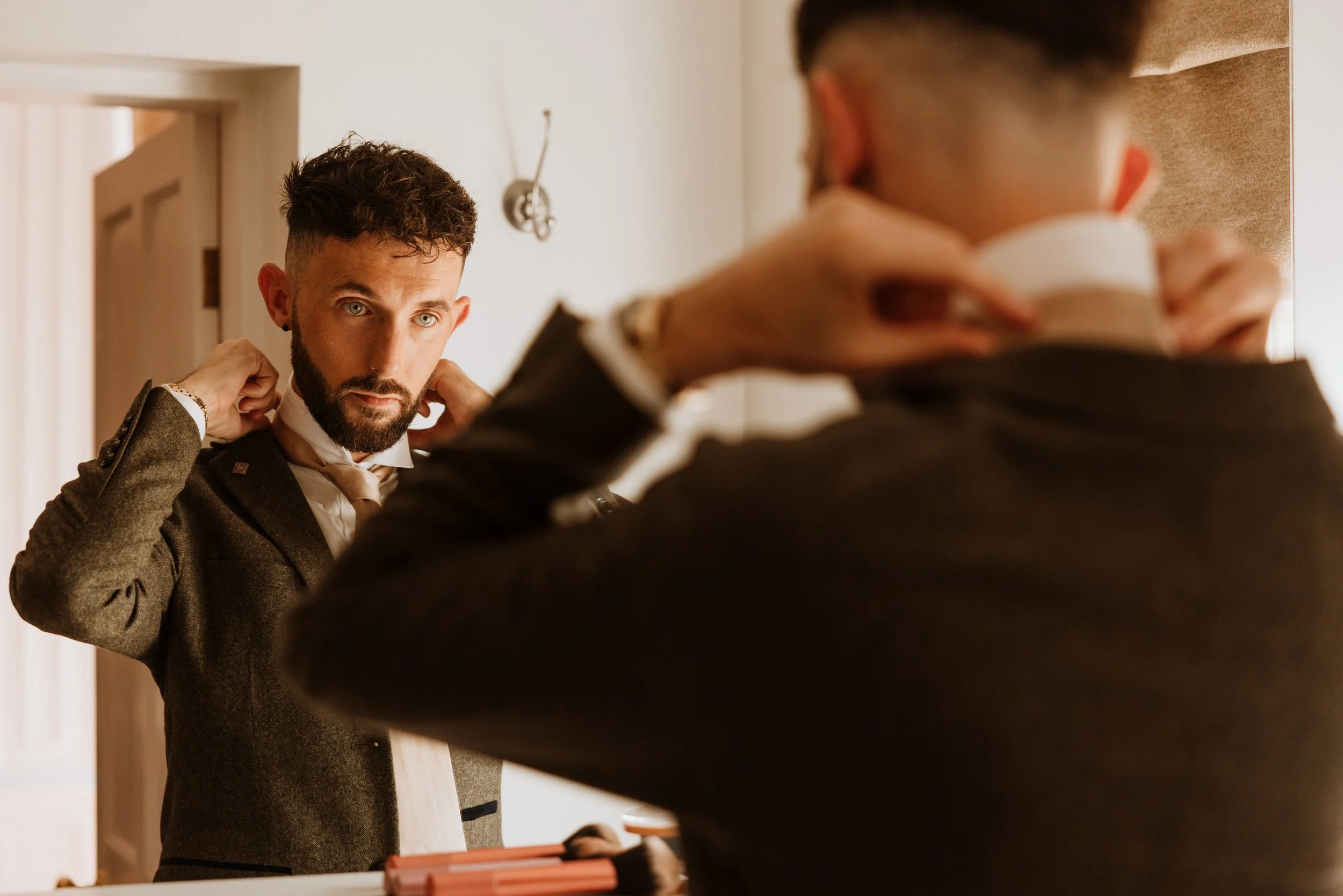 Man adjusting his suit in front of a mirror as another person holds a cup nearby.