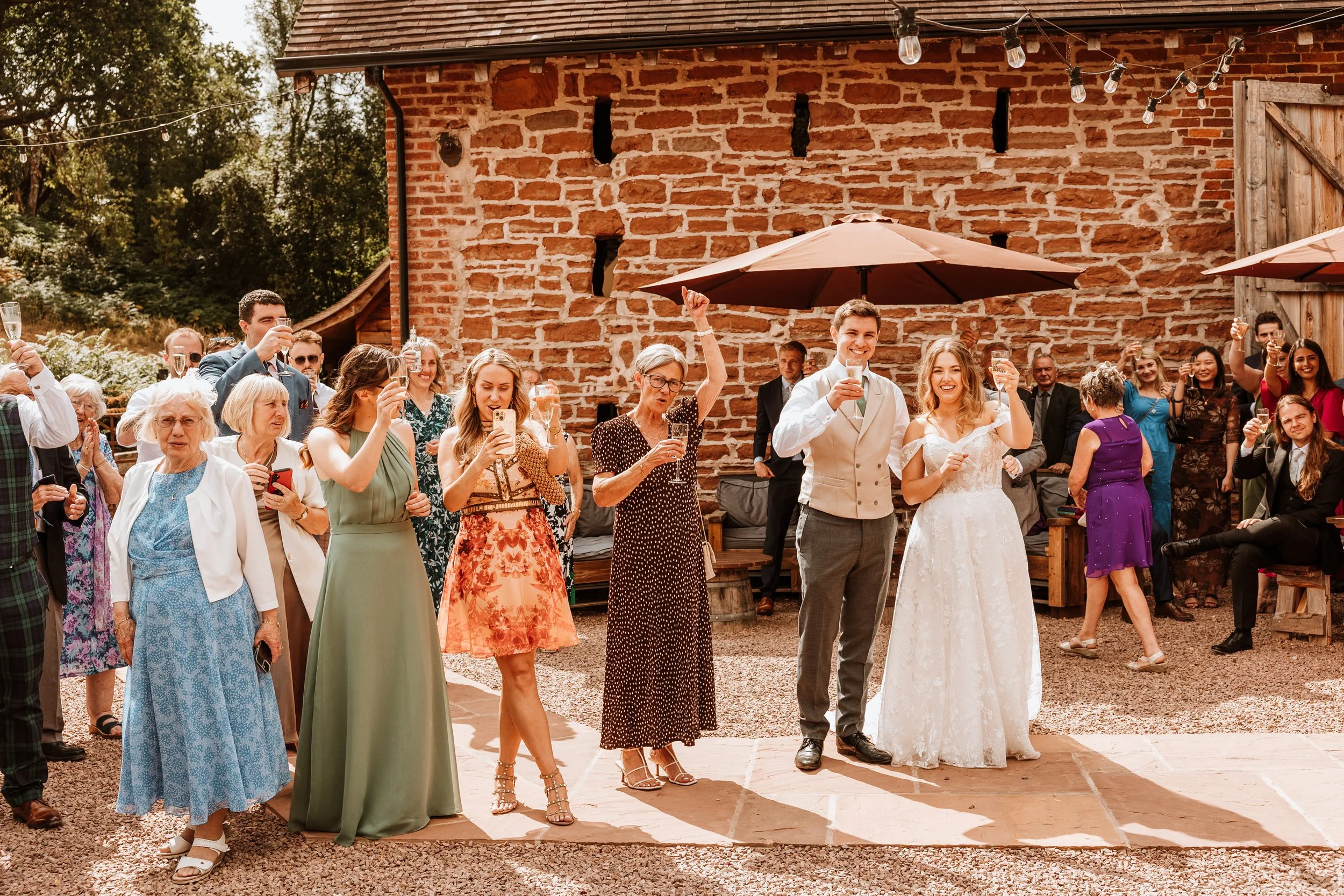 Group of wedding guests raising glasses for a toast outside a rustic brick building with string lights and umbrellas.