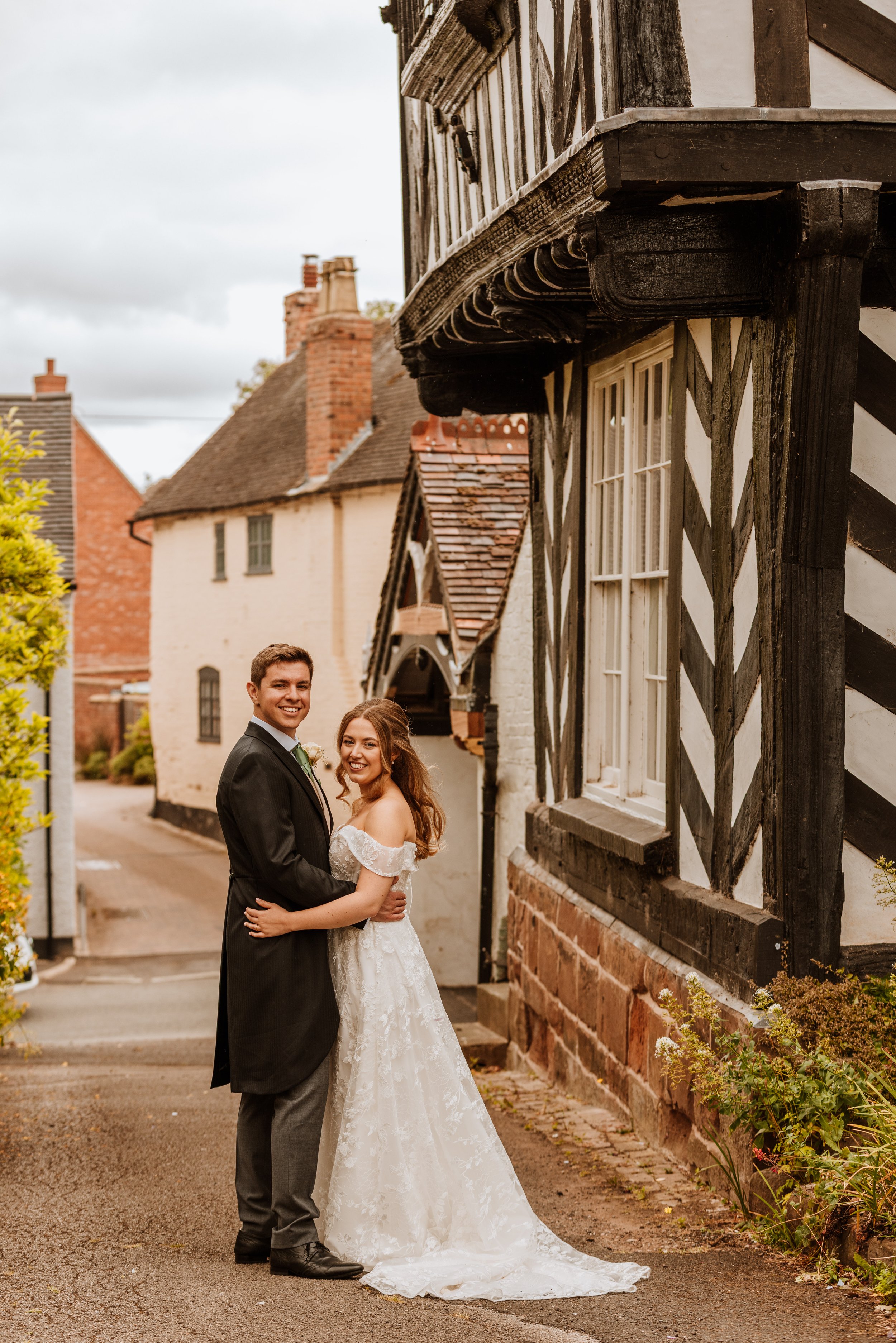A newlywed couple in wedding attire standing on a street in front of old, historic buildings with black and white timber framing, smiling and embracing each other.