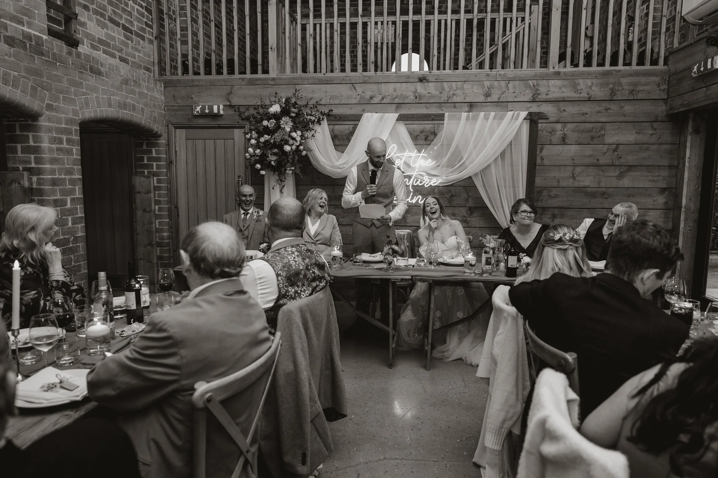 A wedding reception with a bride and groom at a head table, with guests seated facing them, and a person giving a speech at a microphone in front of a floral and draped backdrop in a rustic venue.