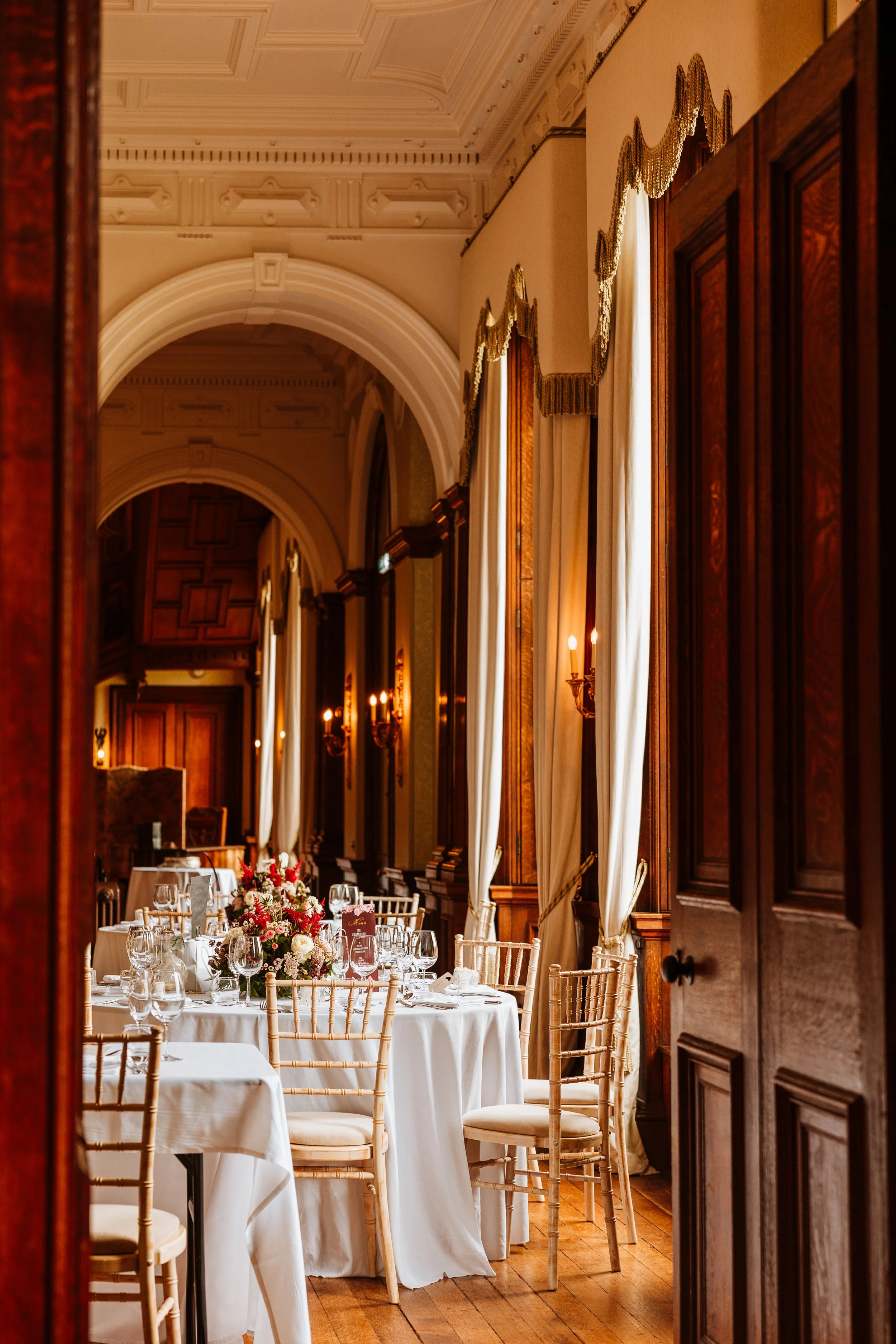 Elegant dining room with round tables covered in white tablecloths, decorated with floral centerpieces, set with glassware and chairs, large windows with curtains, wood paneling, and warm lighting.