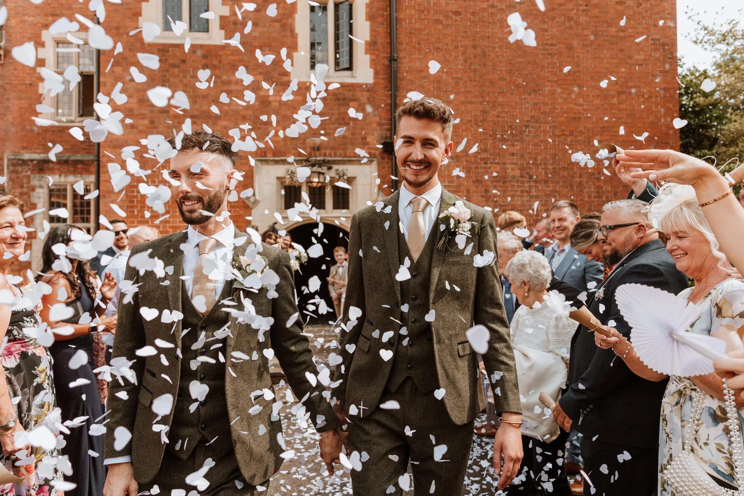 Two men in suits walking hand in hand through a shower of white confetti in front of a red-brick building, surrounded by smiling guests at a wedding celebration.