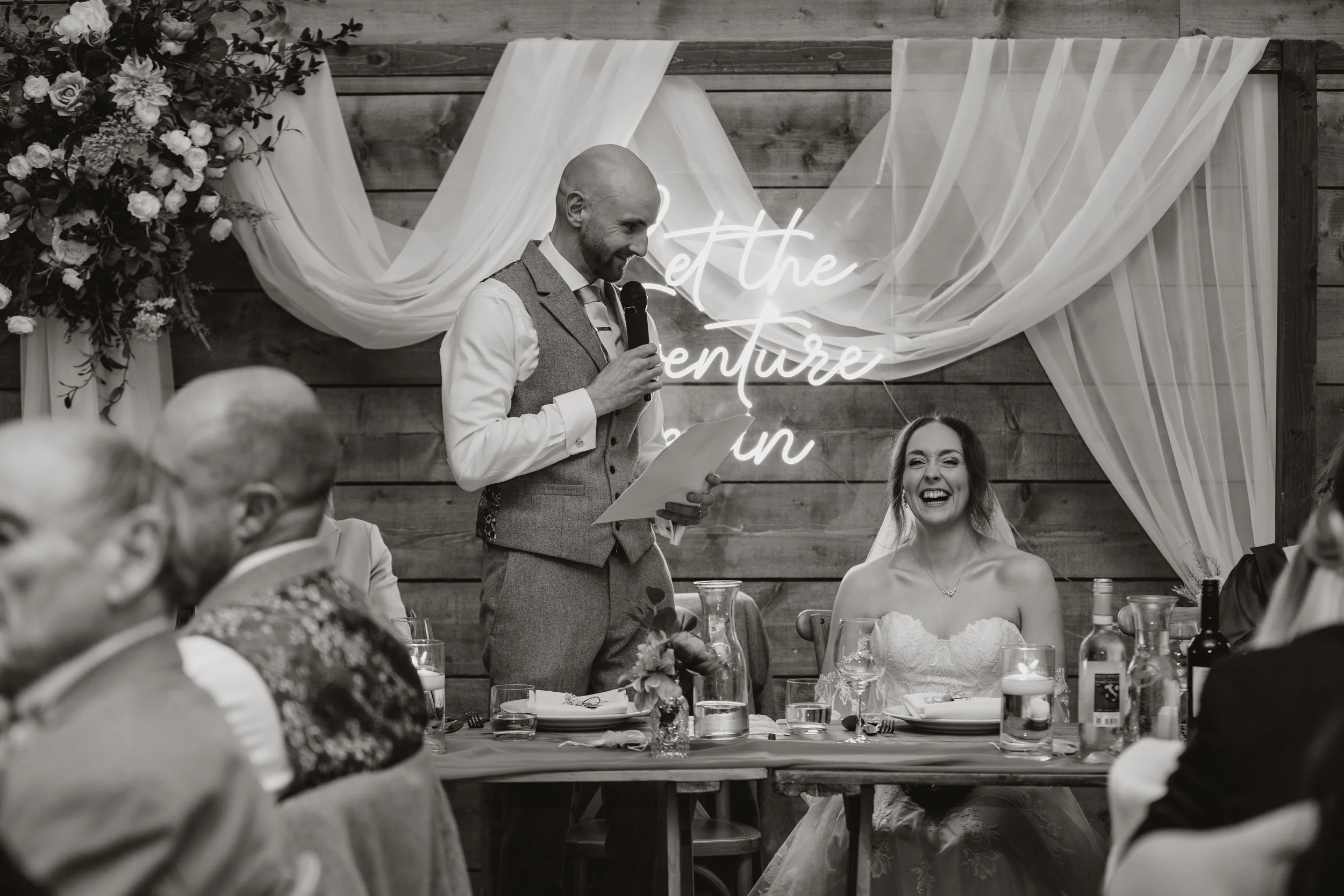 A wedding reception with a man giving a speech while a bride laughs. The scene is decorated with drapery, flowers, and a neon sign in the background.