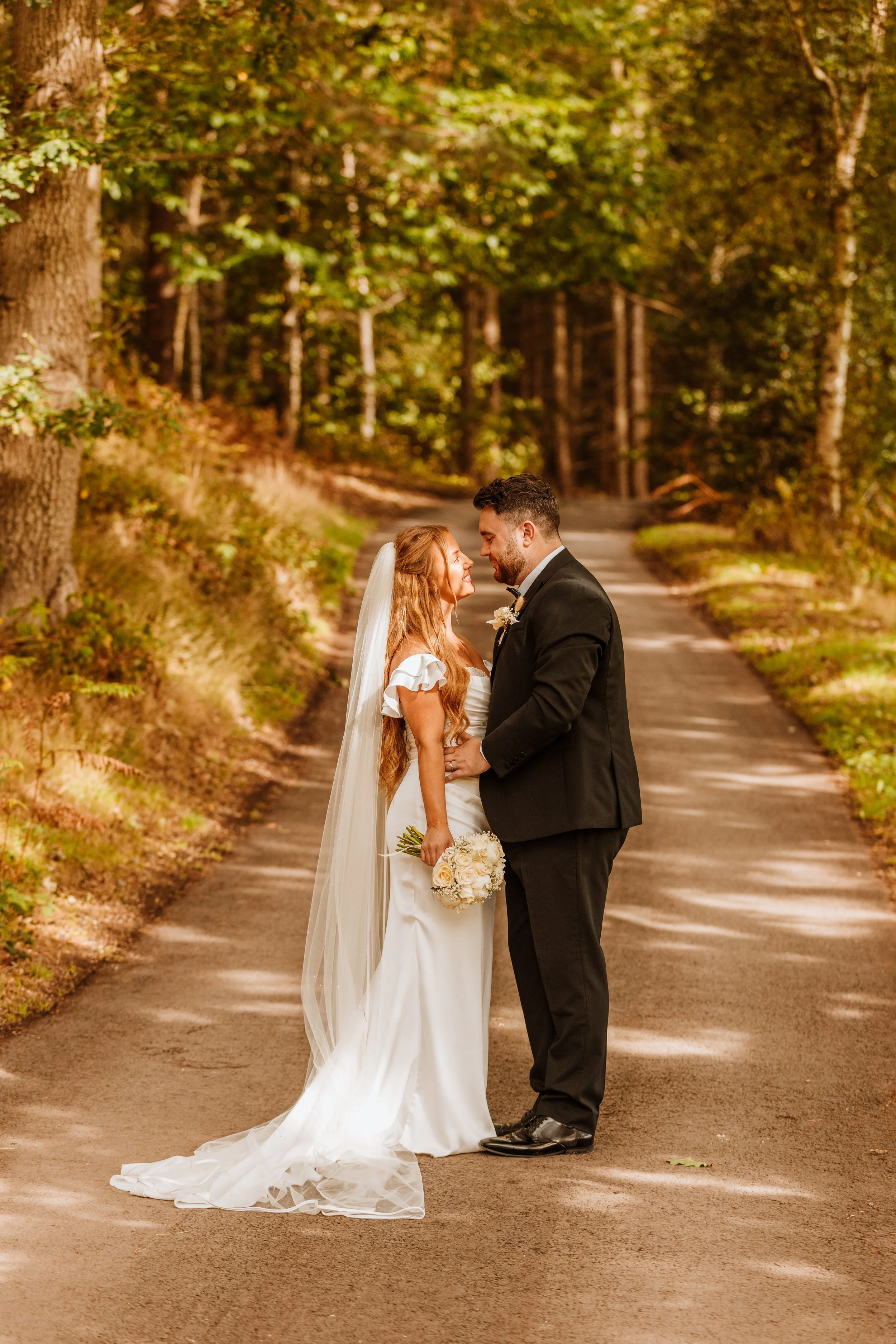 A bride and groom standing close together on a forest path during their wedding, smiling at each other, with the bride holding a bouquet of white roses.