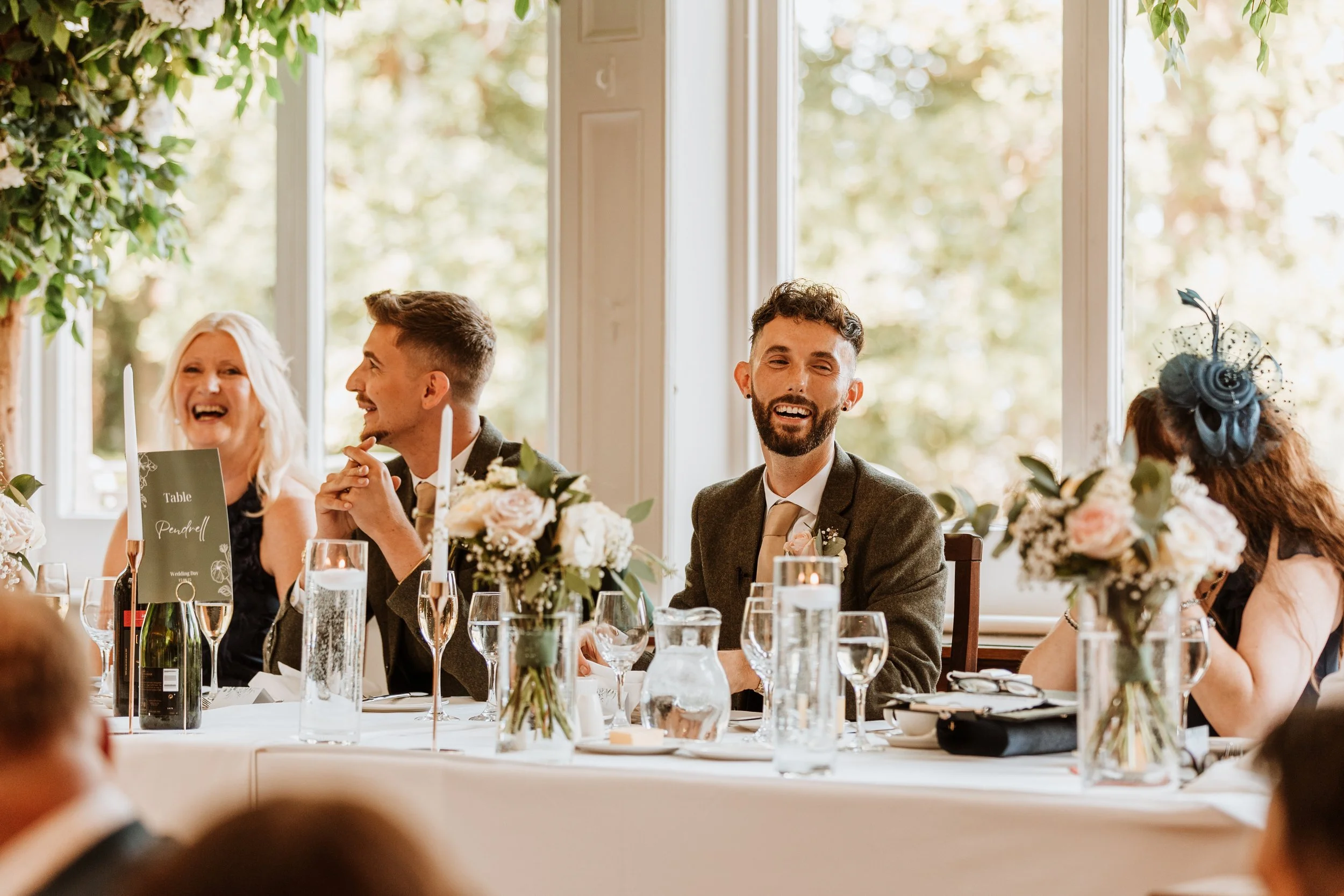 People sitting at a decorated table, smiling and talking, during a celebration in a bright room with large windows and greenery outside.