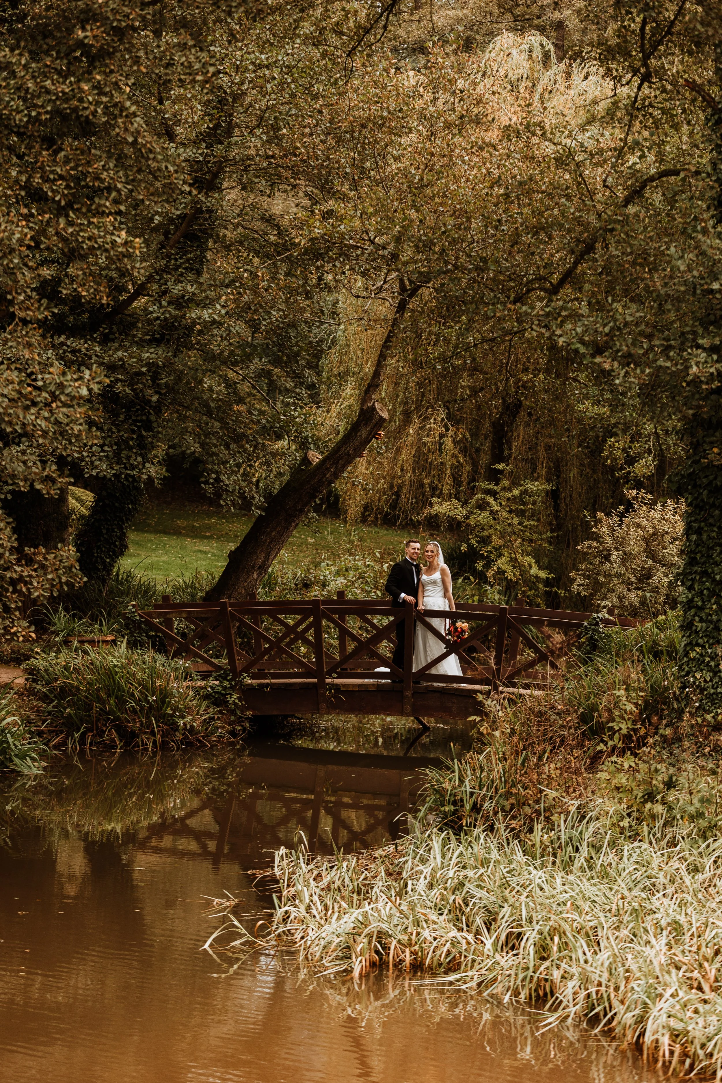 A bride and groom standing on a wooden bridge over a small pond in a forested area during daytime, surrounded by autumn foliage.