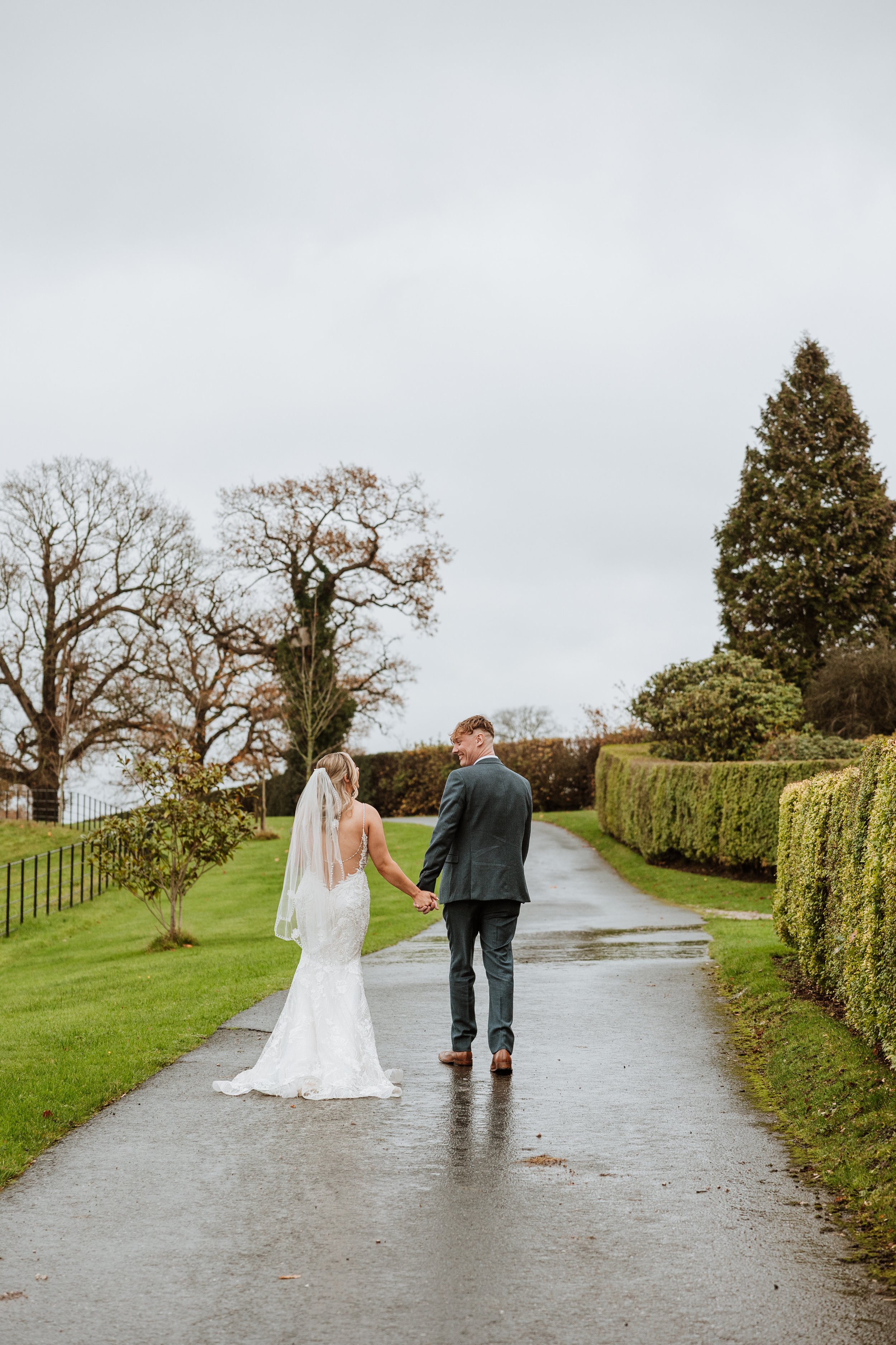 A bride and groom walk hand in hand down a wet, paved path in an outdoor setting with overcast sky and lush green hedges lining the path at Grange Barn, Cheshire.
