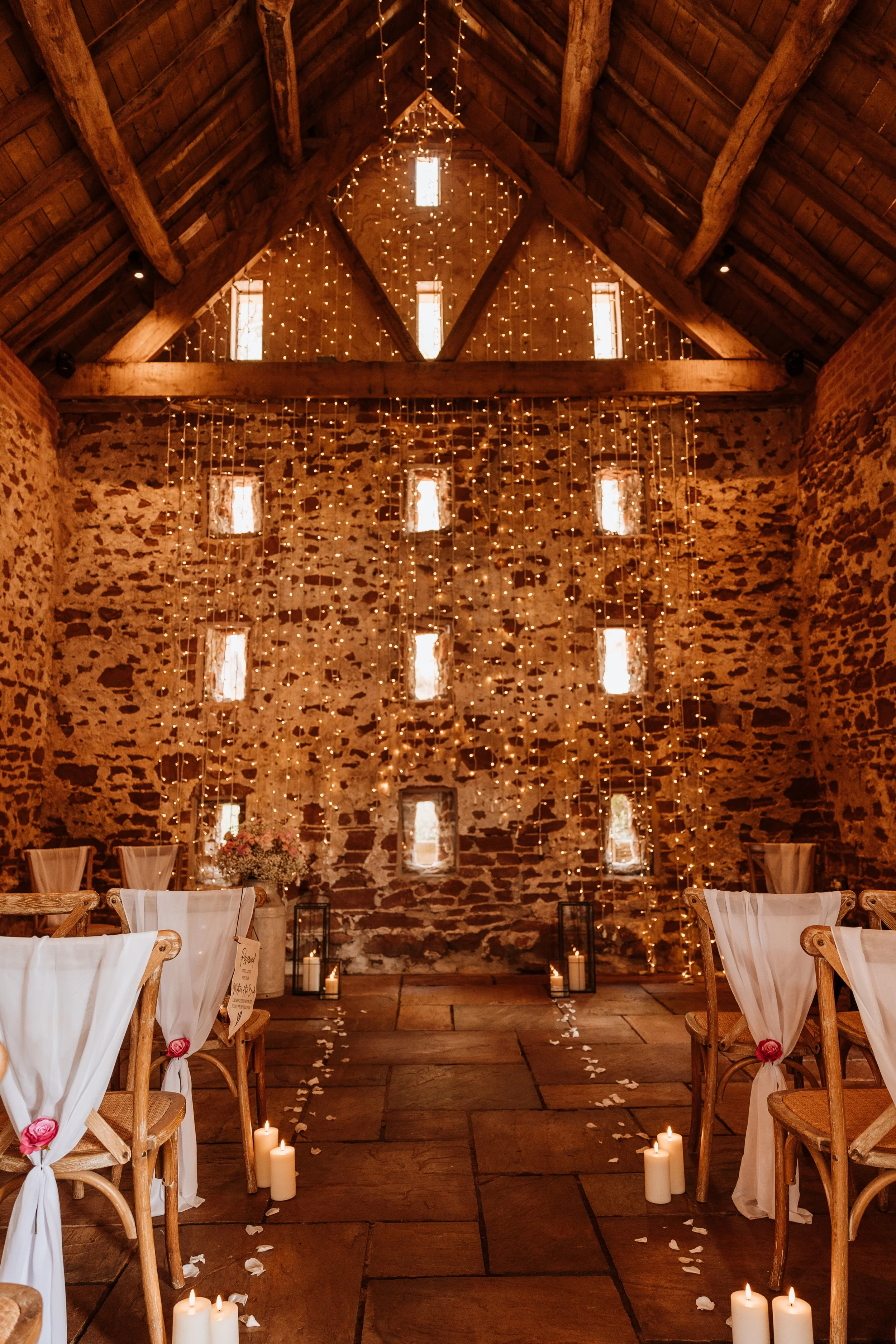 Wedding ceremony setup in a rustic barn with candles, string lights, and chairs decorated with white fabric and pink flowers.