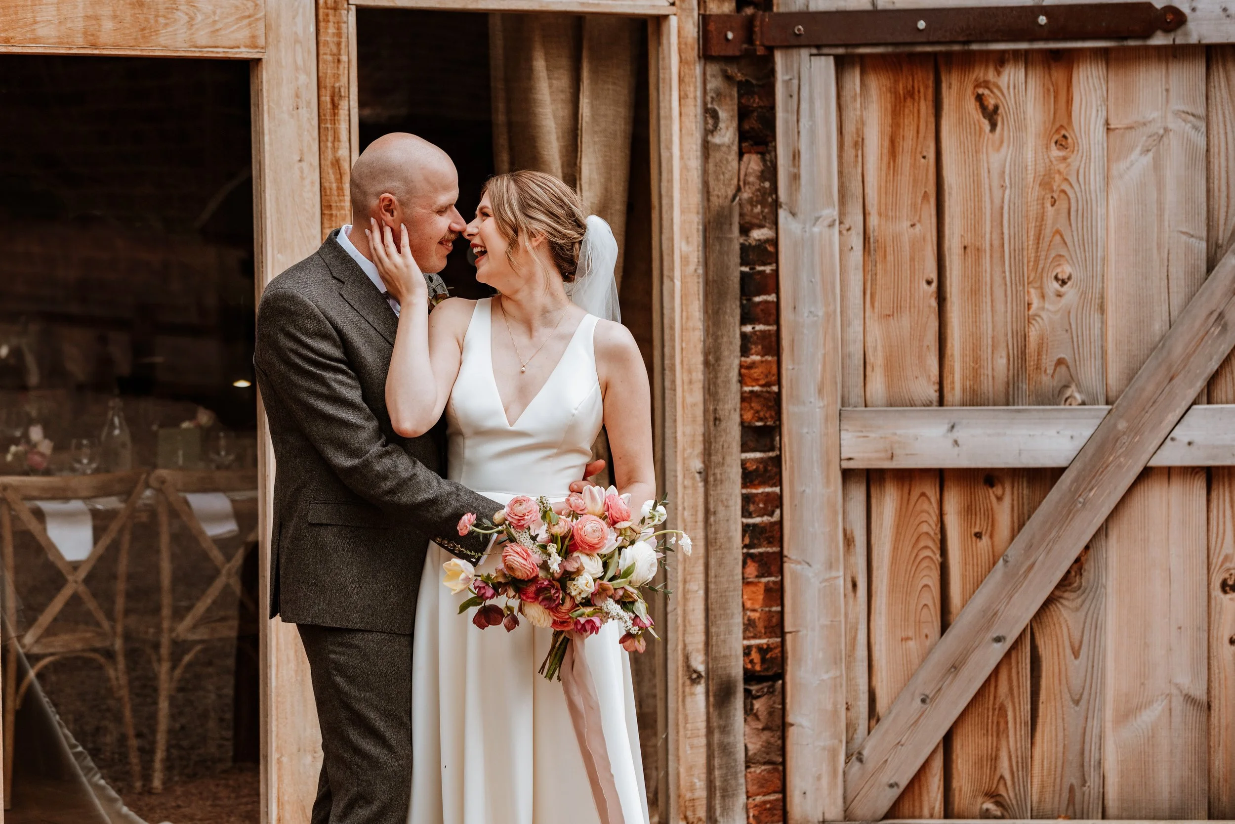 A bride and groom sharing a kiss inside a rustic barn, with the bride holding a bouquet of pink and white flowers.