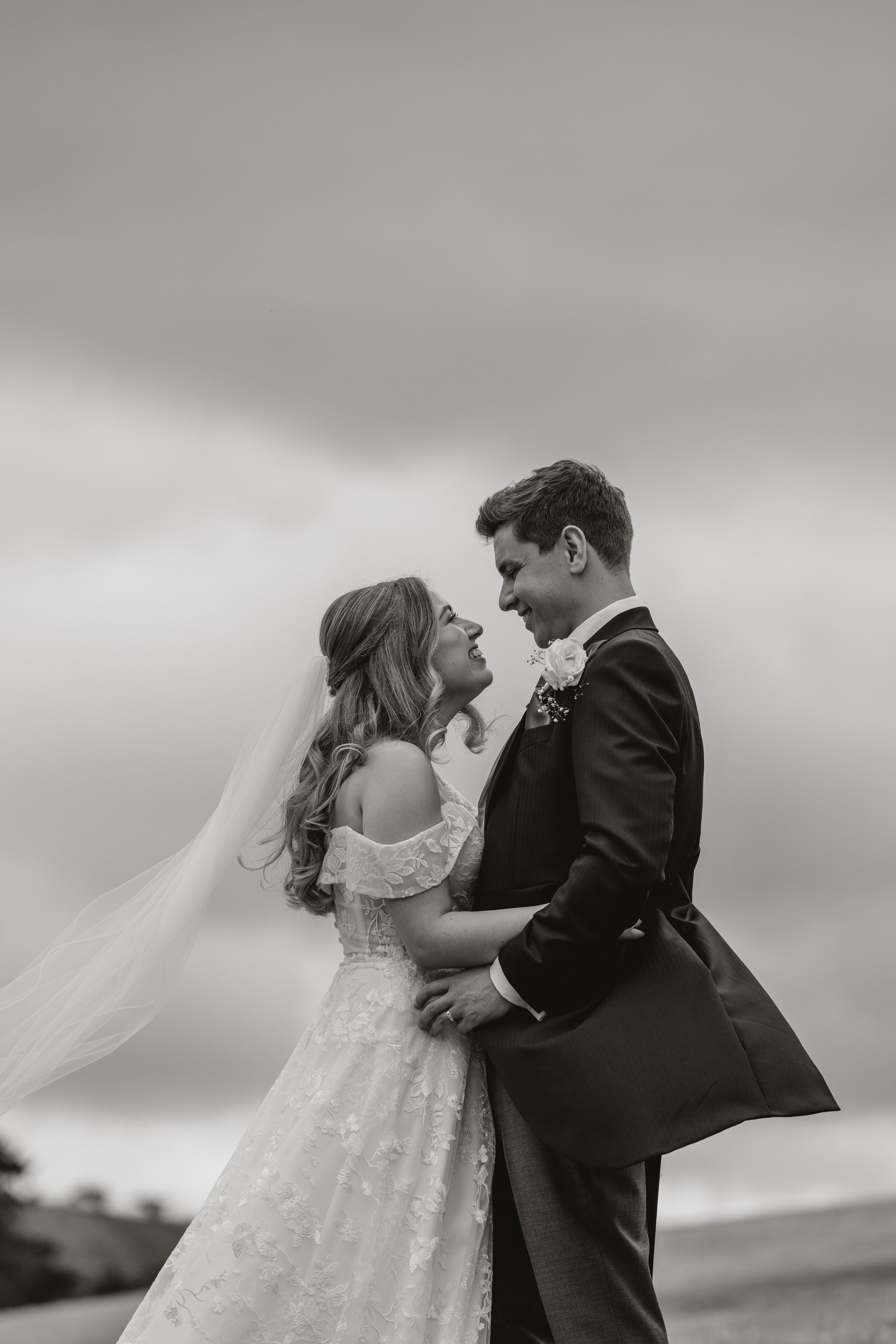 Black and white photo of a bride and groom smiling and embracing outdoors on a cloudy day.