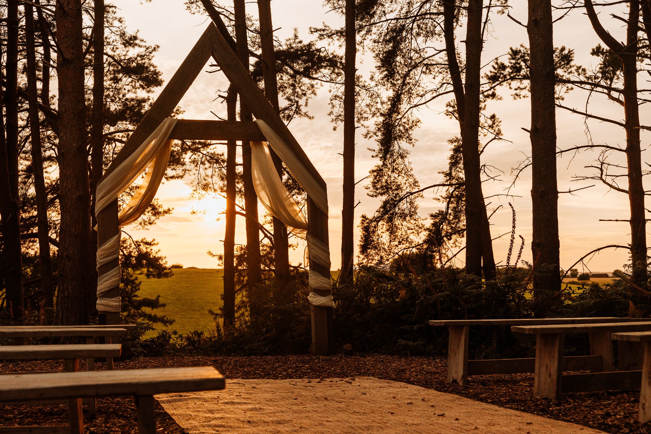 A wooden wedding arch decorated with cream-colored fabric, set outdoors among tall trees with a sunset sky in the background. Bridal Barn, Claverley, Bridgnorth, Shropshire. 