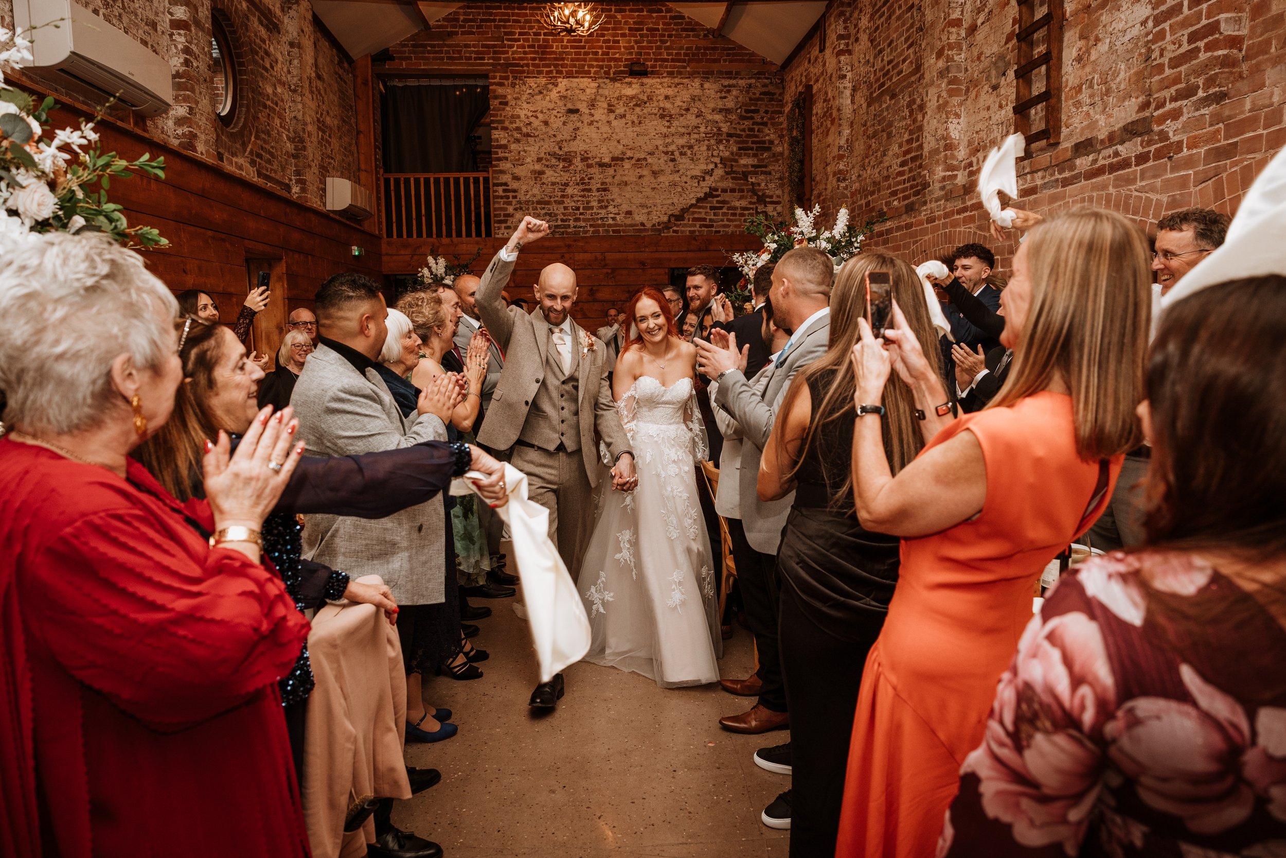 A bride and groom walking hand-in-hand through a crowd of cheering guests at a wedding reception in a rustic brick venue.
