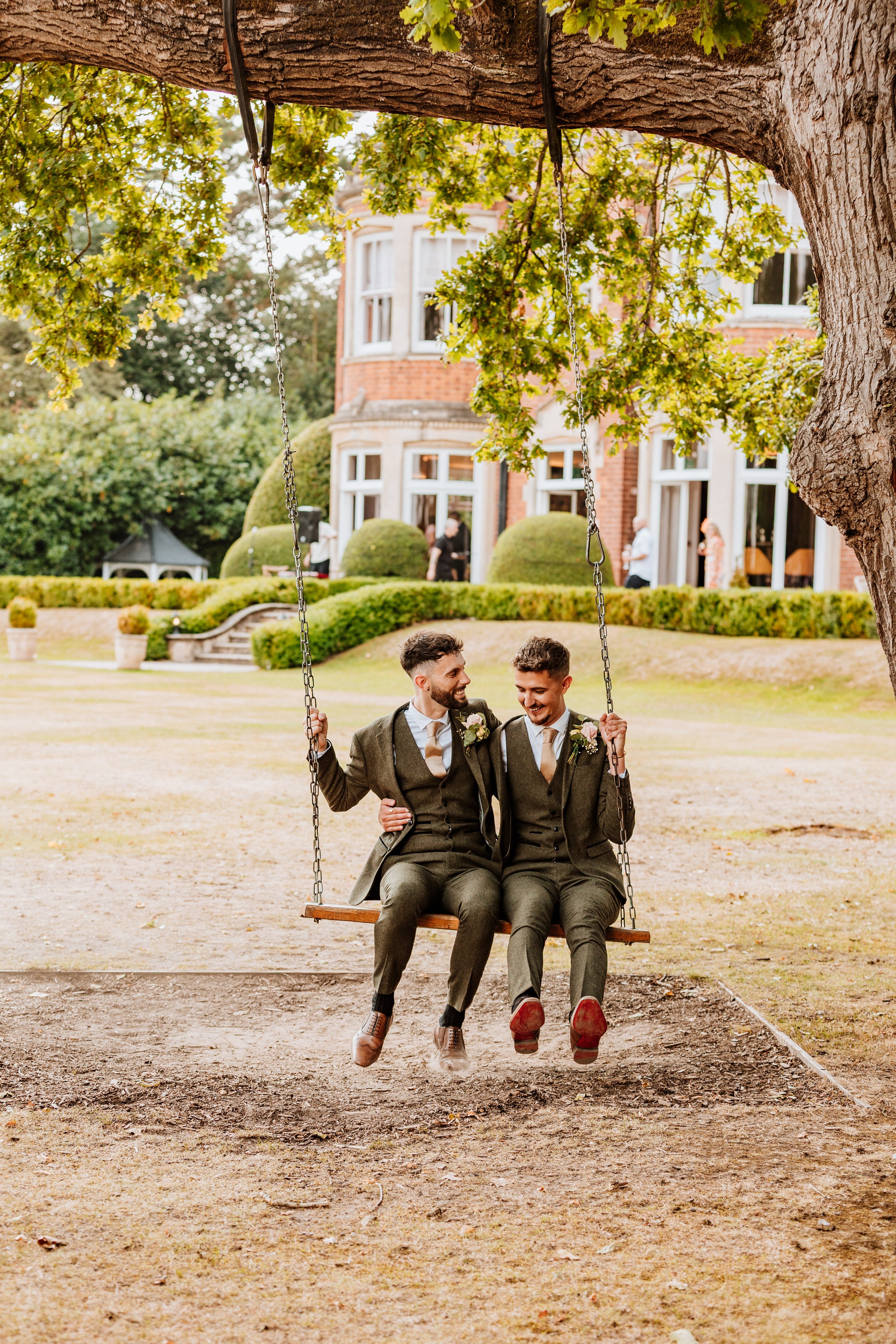 Two men in suits sitting on a swing hanging from a tree in a garden, smiling and talking. Large house and guests in the background.