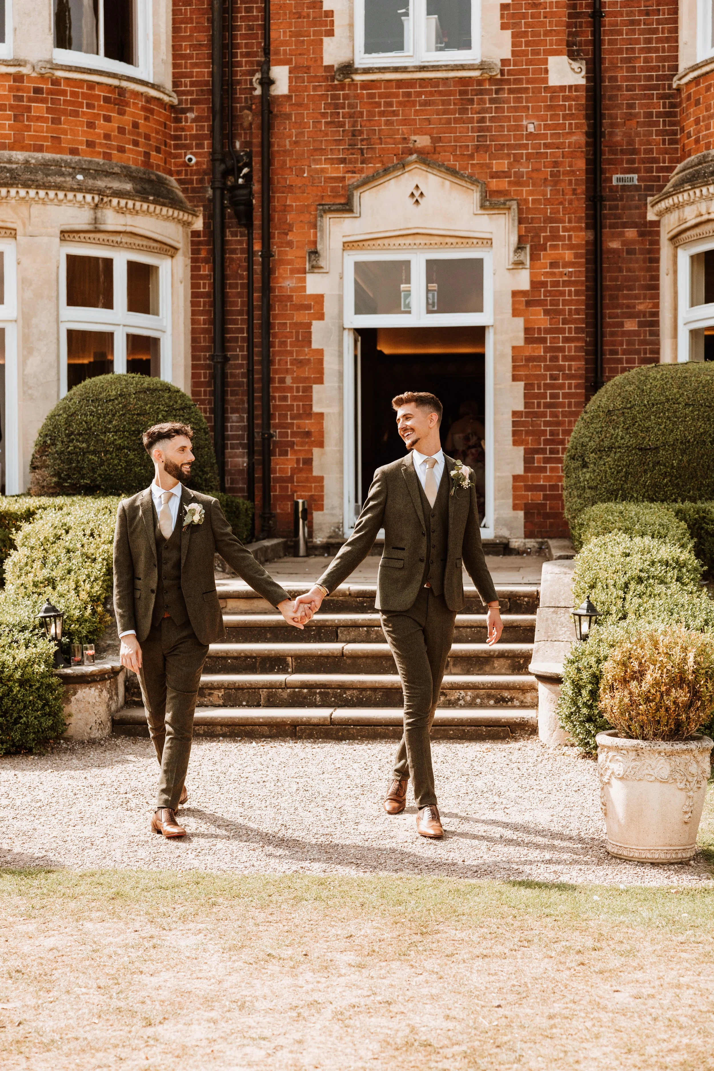Two men in suits holding hands and walking outside a brick building with stairs, surrounded by greenery, smiling at each other.